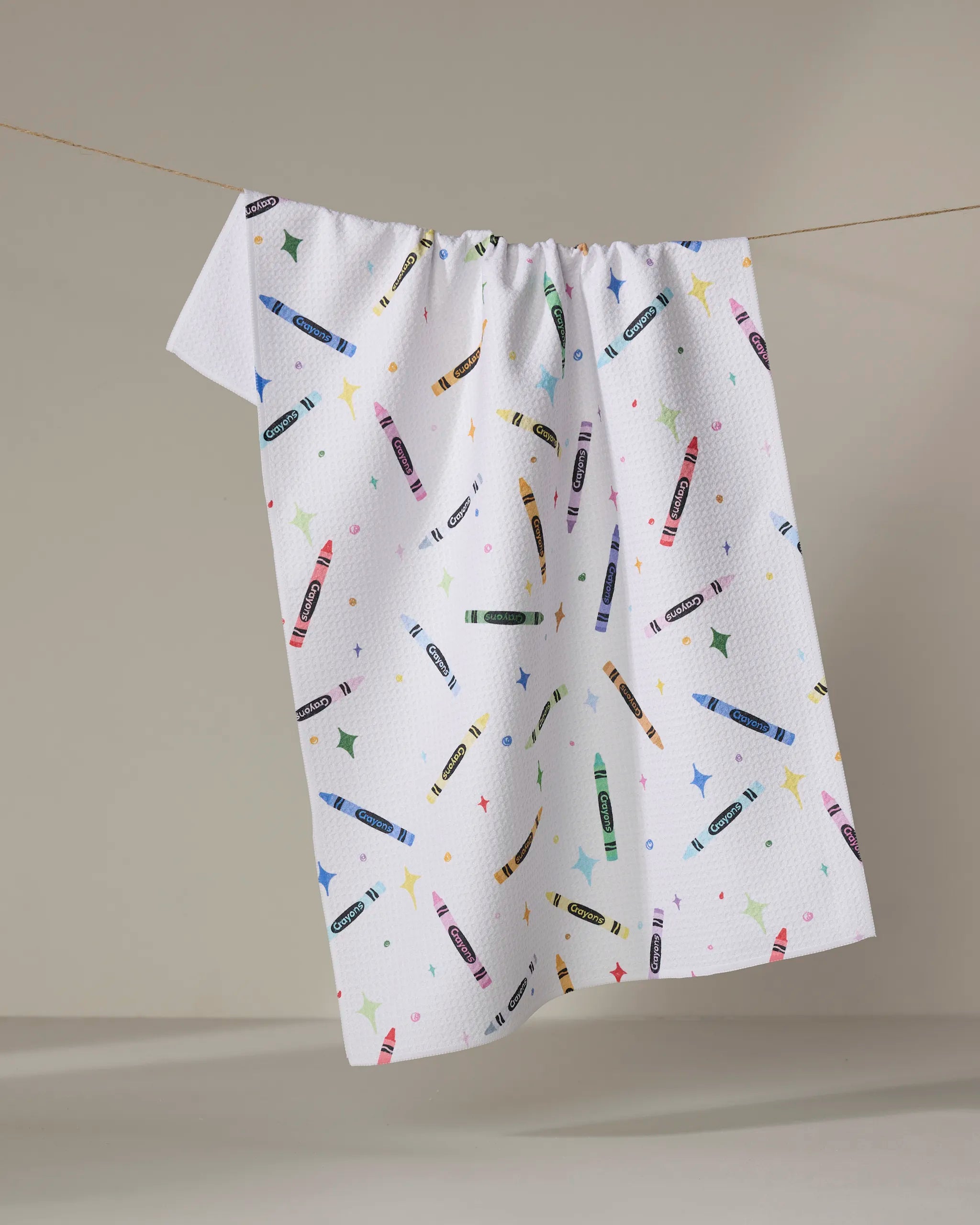 The Vaishali Design Studio "Colored Crayons" tea towel, featuring colorful crayon and star designs, quick-dries and hangs on a thin clothesline against a plain, neutral background.