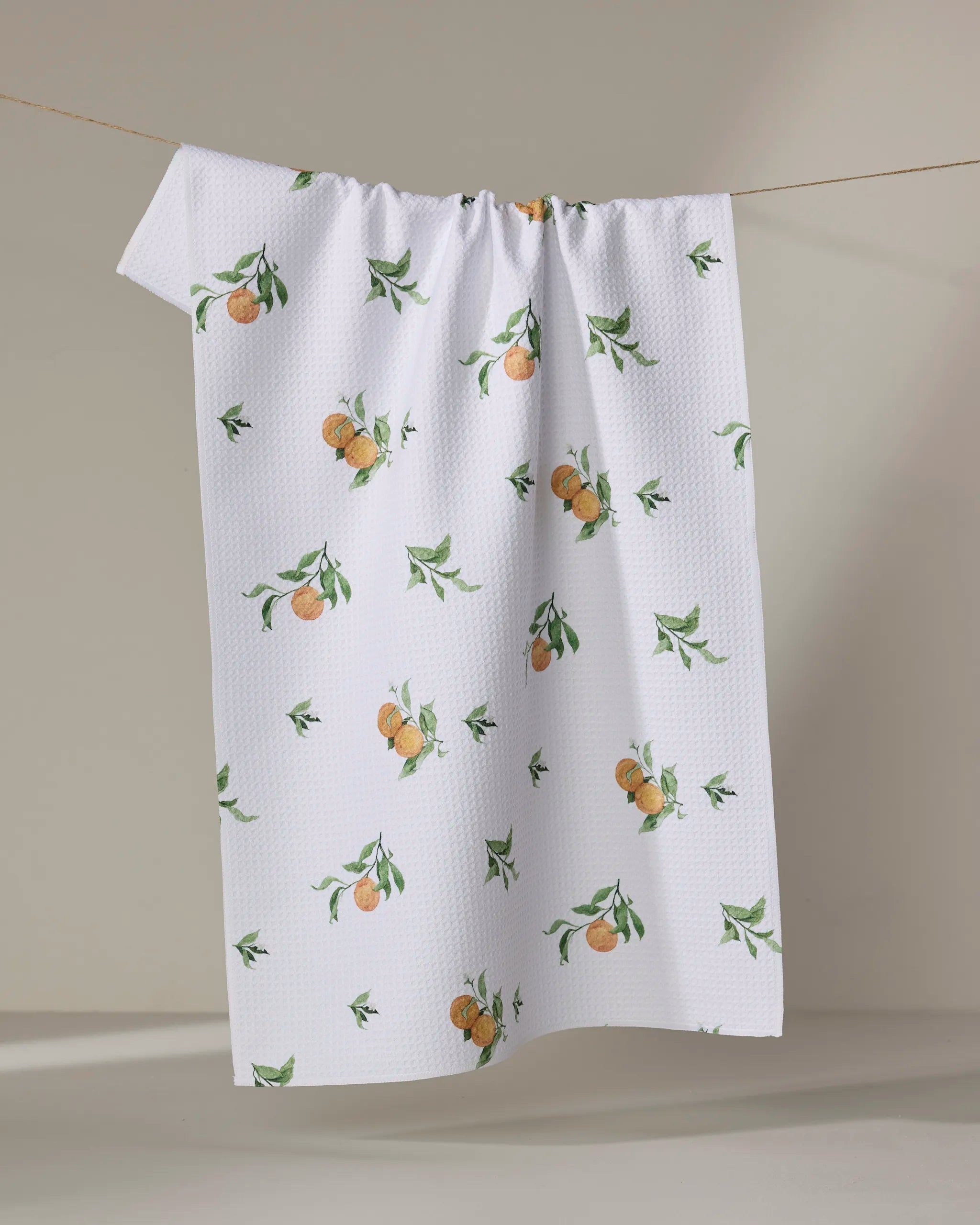 The Aubrey Fairchild Comfort Fruit tea towel, featuring a woven orange fruit and green leaf pattern and made from recycled materials, hangs on a beige background.