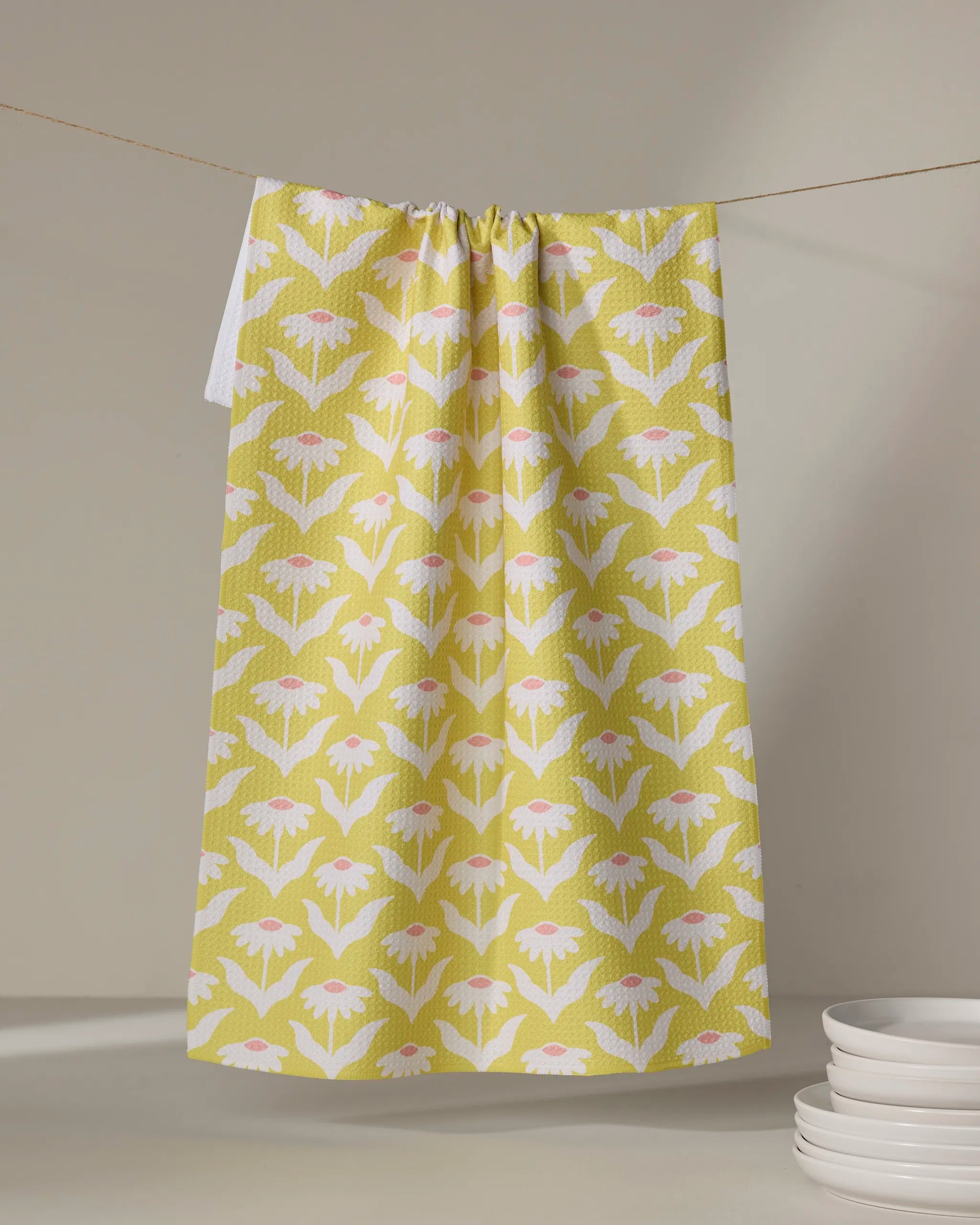 The Coneflower recycled kitchen towel by Kayla Ann, featuring a yellow base with pink and white floral patterns, hangs on a string beside a stack of white plates on a light-colored surface.