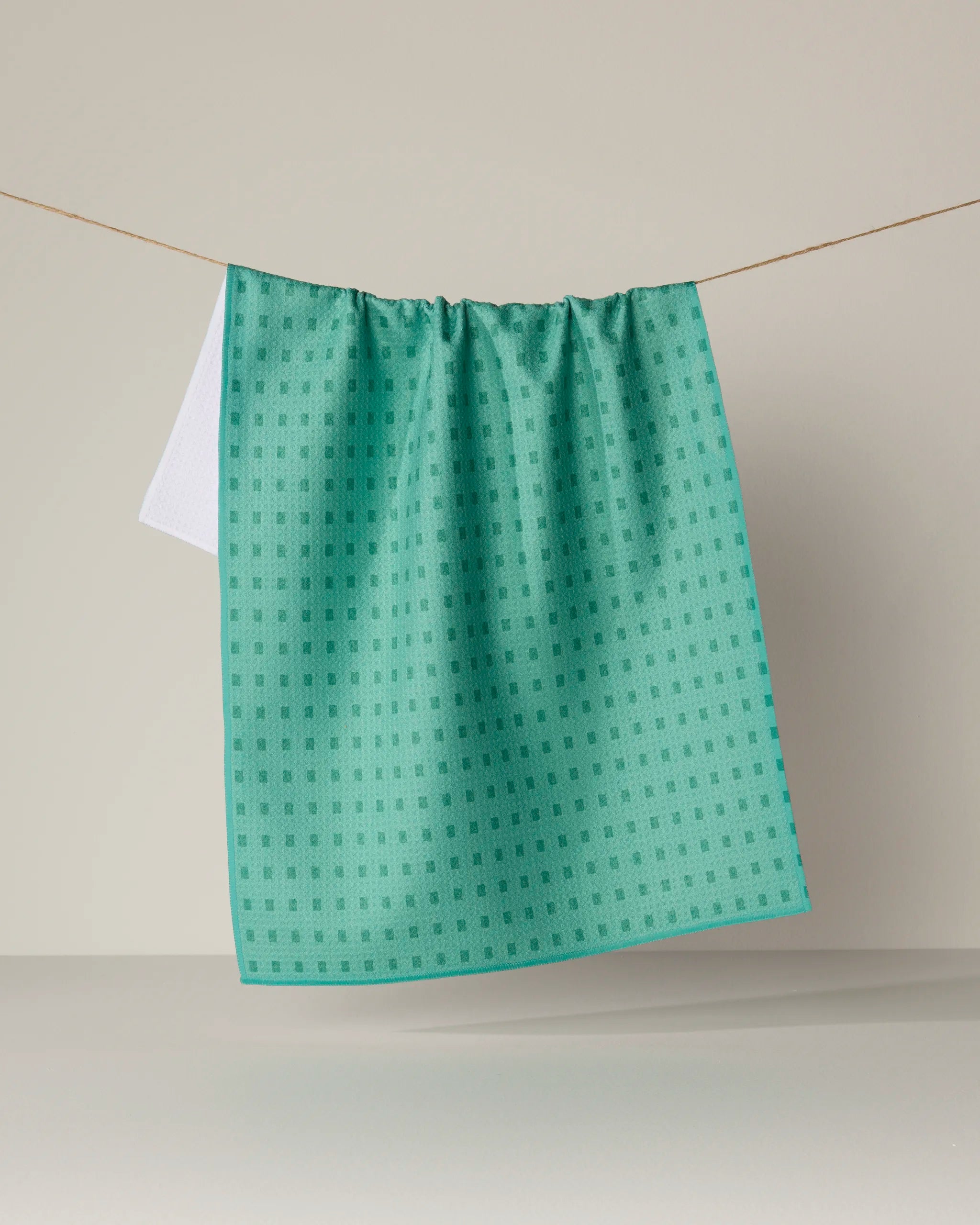 A Geometry Confetti Tiles kitchen towel in a teal pattern hangs on a string against a neutral background, with a small section of a white towel visible behind it.