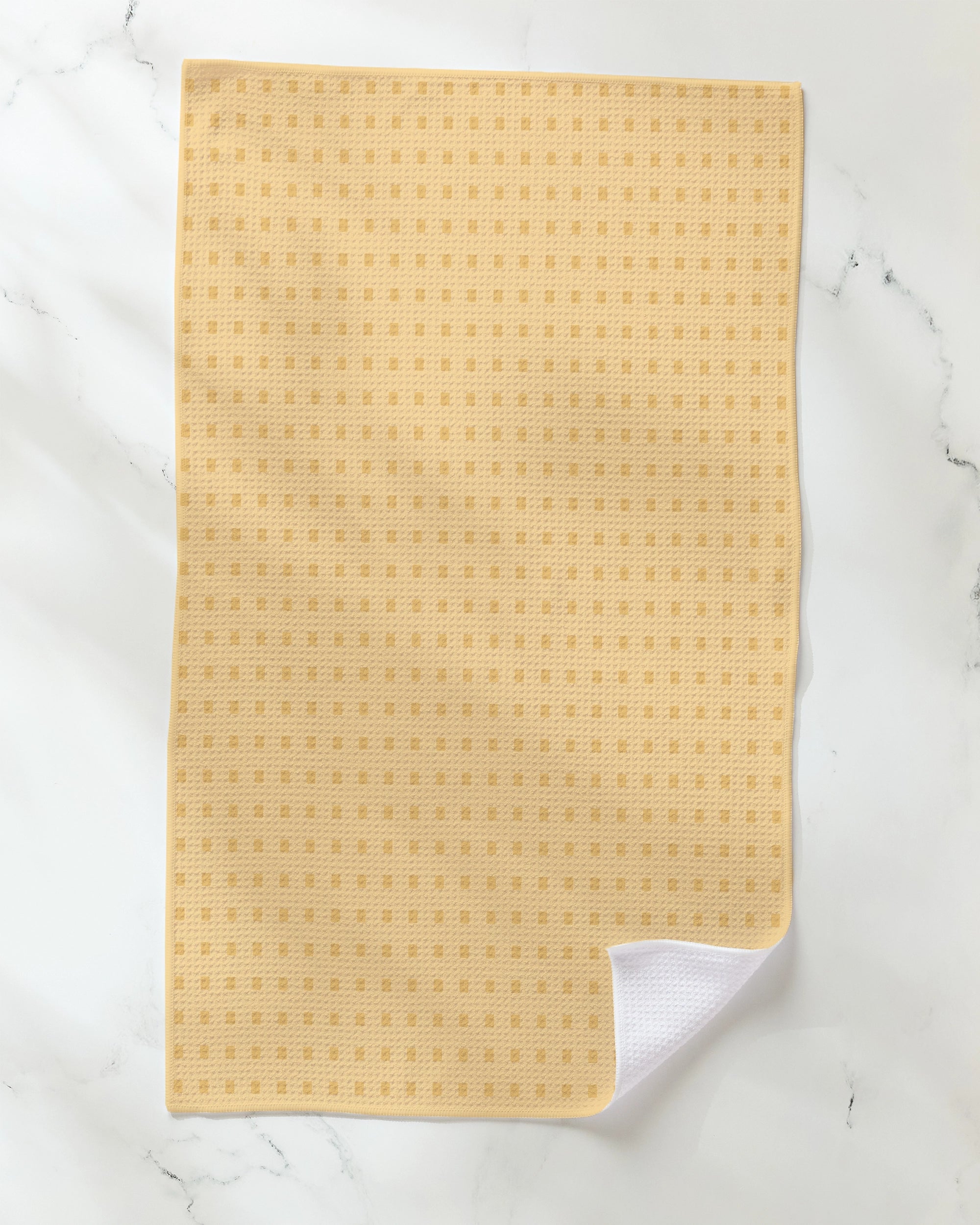 The Geometry Confetti Tiles tea towel, featuring a light yellow hue and a small square check pattern, lies flat on a white marble surface with one corner folded to reveal the white underside.