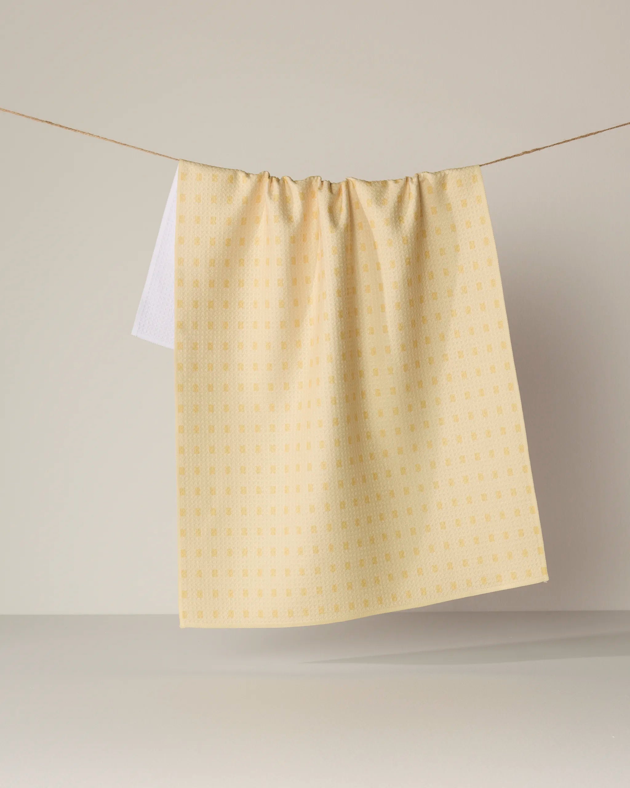 A Confetti Tiles tea towel by Geometry, featuring a yellow-checkered pattern, hangs neatly on a clothesline against a plain, light gray background.