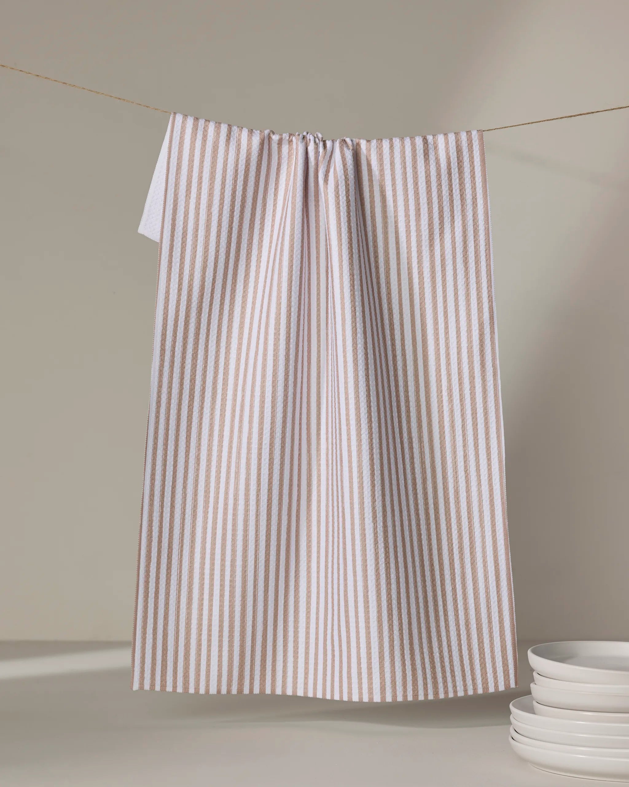 The Kailee Wright "Cookie" kitchen towel, featuring beige and white vertical stripes, hangs on a string against a neutral backdrop, with a small stack of white plates in the lower right corner.