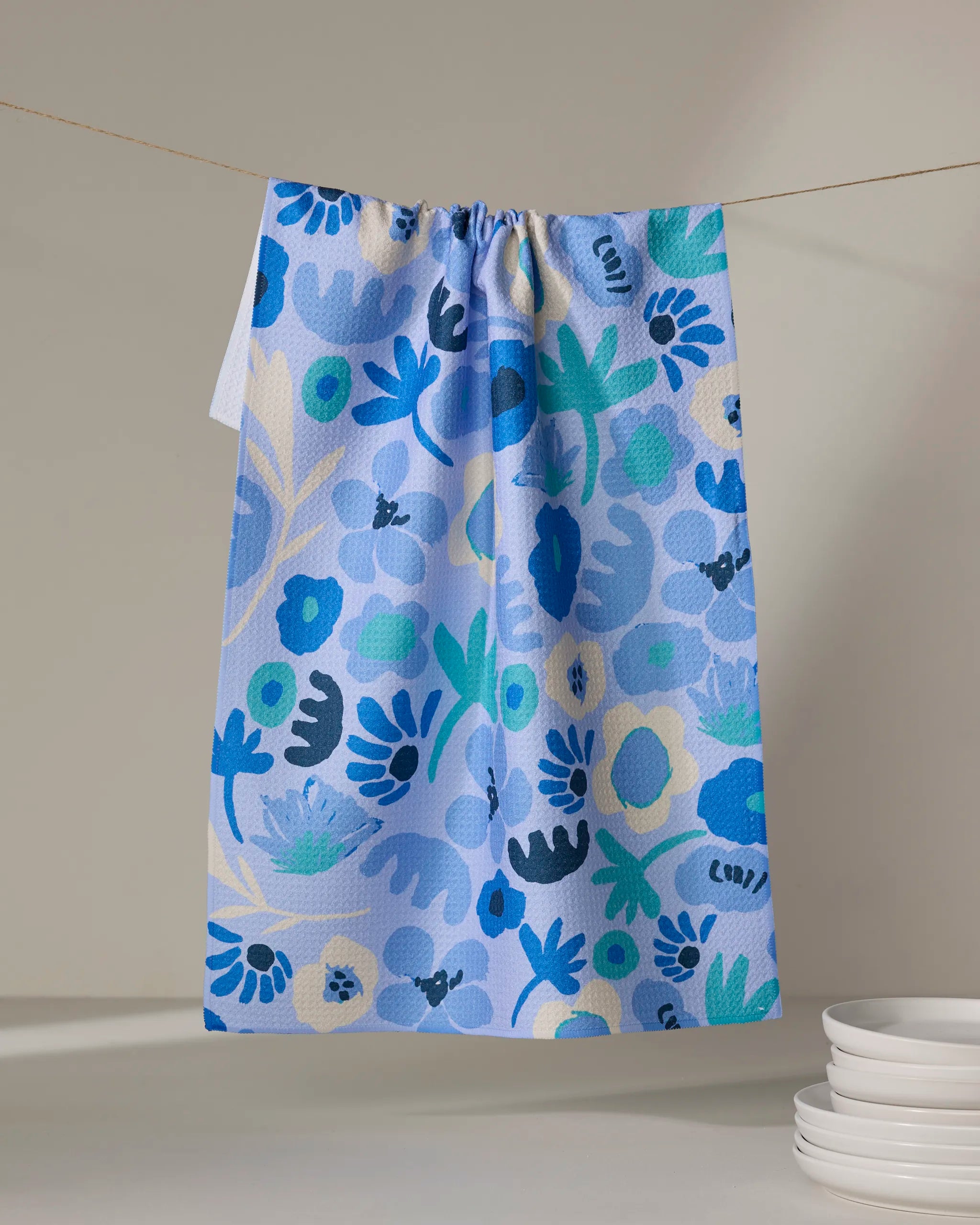 The Cool Blooms tea towel by Sareka Unique, featuring a bold blue, green, and cream floral pattern on light blue recycled fabric, hangs on a line beside stacked white plates resting on a neutral surface.