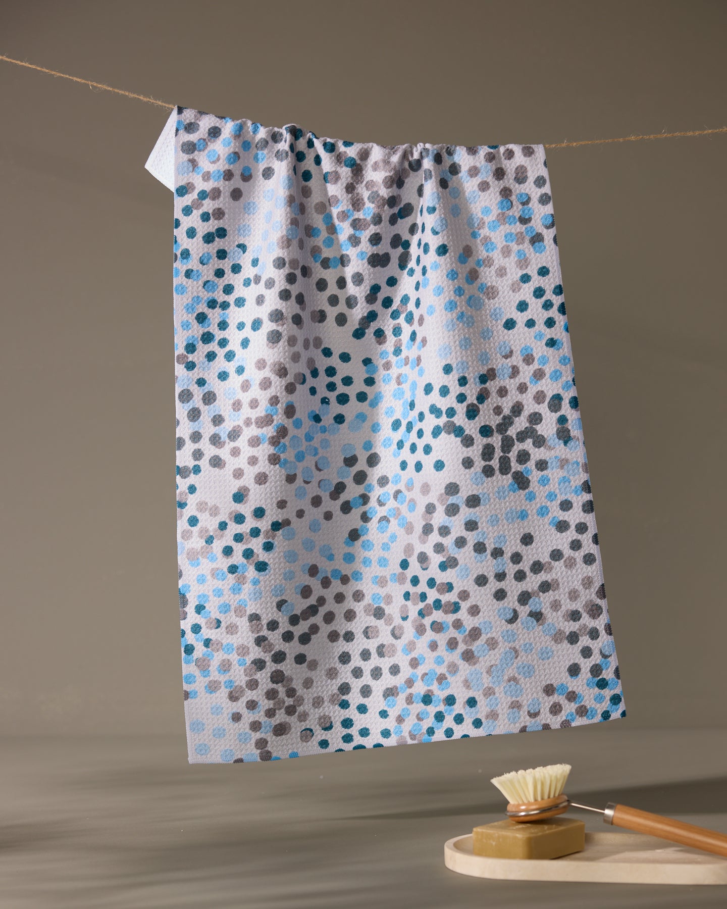 The Geometry Cool Dots ultra-absorbent white kitchen towel with blue, gray, and black dots hangs on a clothesline above a wooden brush with light bristles resting on a round soap dish in a neutral, softly lit setting.