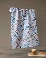 The Geometry Cool Dots ultra-absorbent white kitchen towel with blue, gray, and black dots hangs on a clothesline above a wooden brush with light bristles resting on a round soap dish in a neutral, softly lit setting.