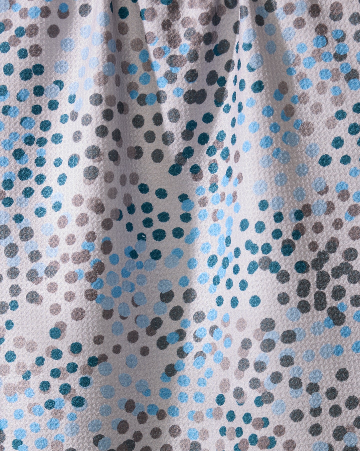 A close-up of Geometry’s Cool Dots kitchen towel shows its textured, ultra-absorbent recycled fabric with an abstract pattern of blue, gray, and black dots scattered on white, highlighted by gentle folds and shadows.