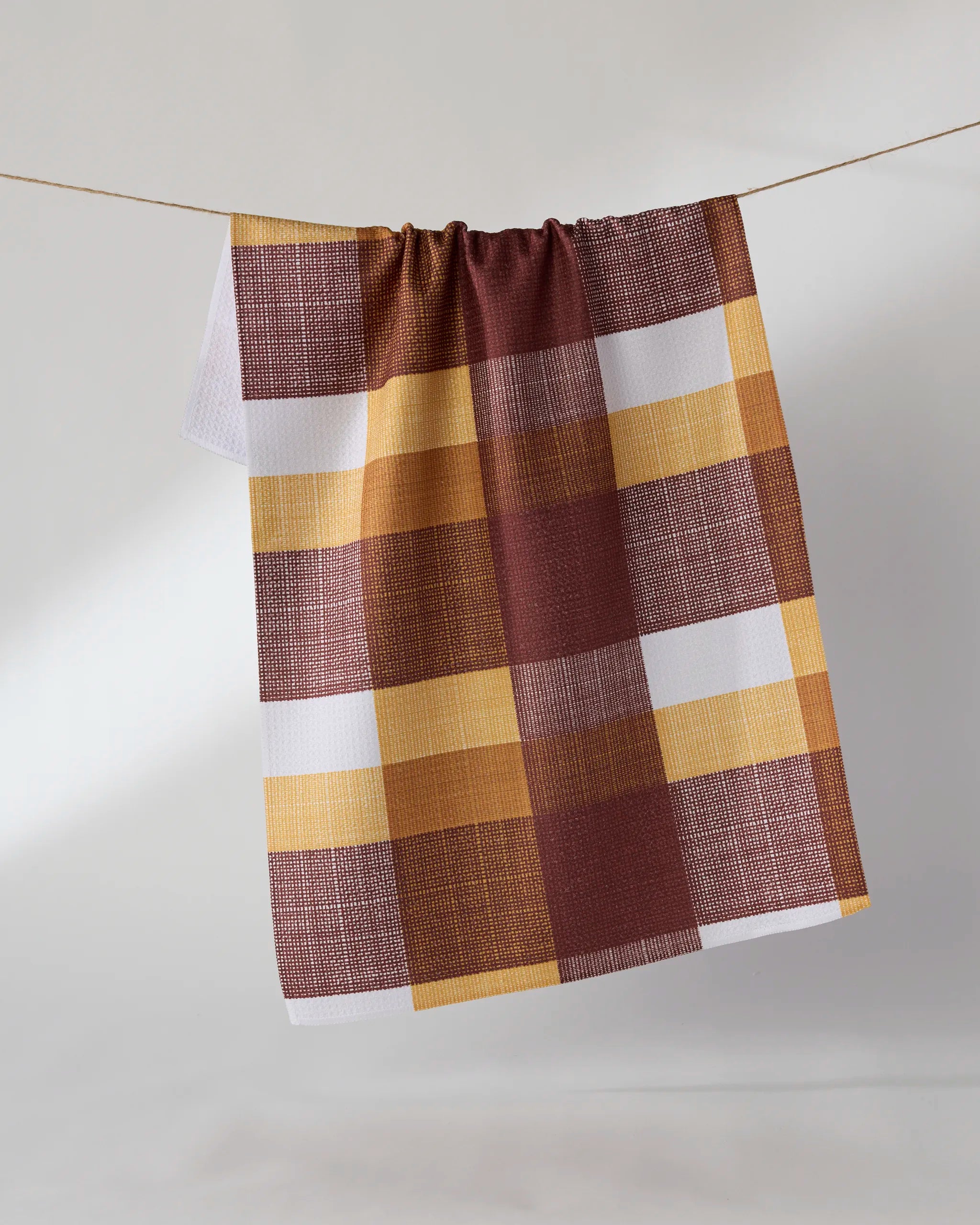 The Geometry Copper Grove kitchen towel, featuring a large brown, yellow, and white plaid pattern and made from recycled materials, hangs on a string against a plain light background.