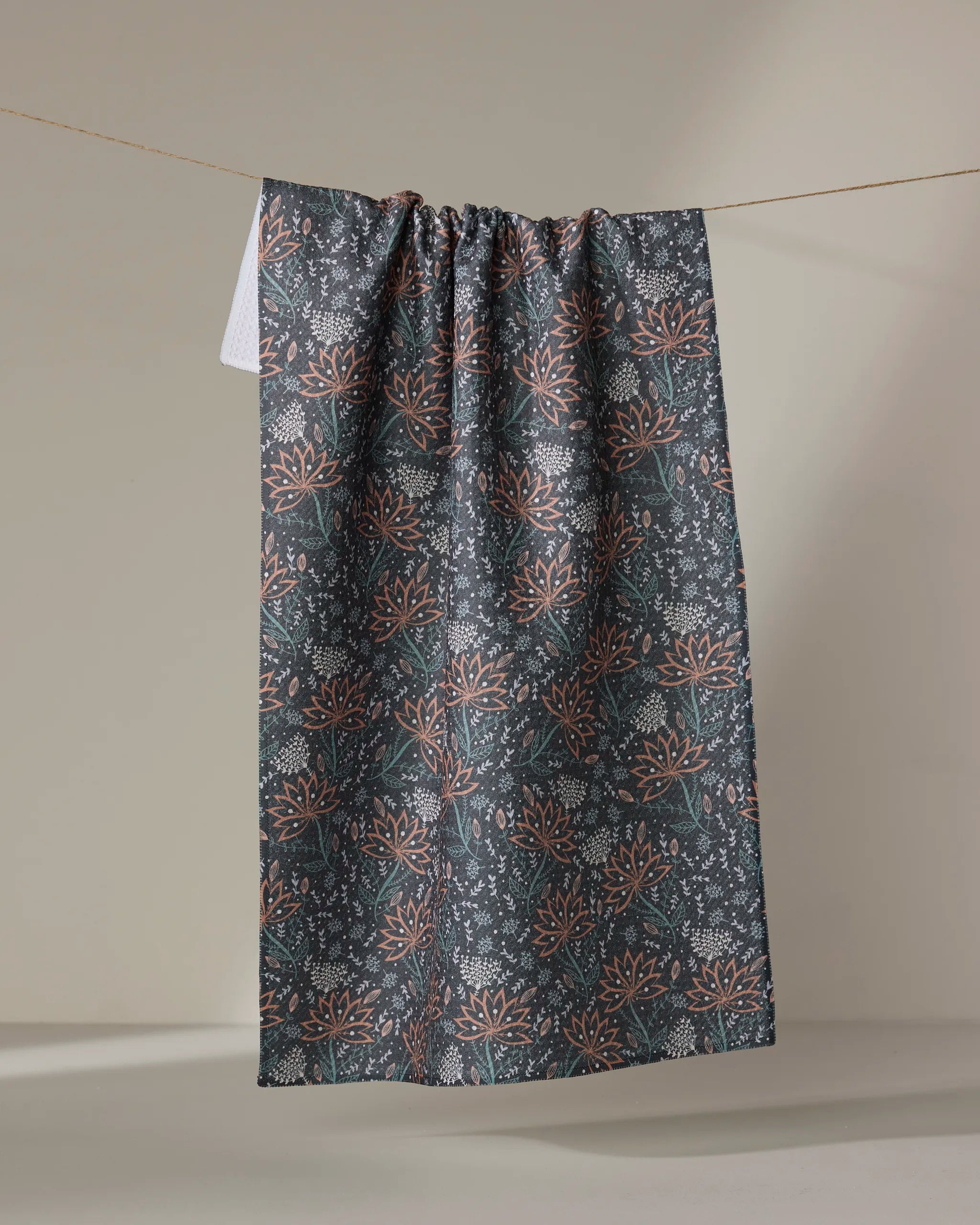 The "Coral Blooms" tea towel by Amita Nair, featuring orange and white floral patterns on dark gray recycled fabric, hangs on a clothesline against a neutral background.