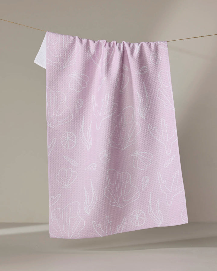 The Coral Cove kitchen towel by Janna Sue Design features light pink, recycled fabric with white sea-inspired line drawings. Absorbent and eco-friendly, it's shown hanging on a clothesline against a neutral background.