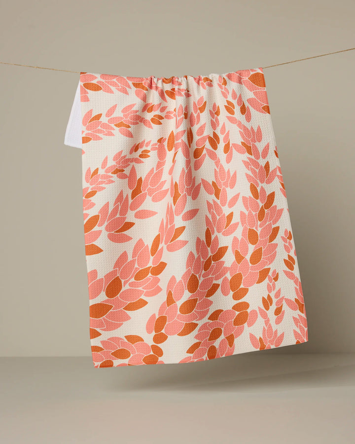 The Geometry Coral Drift tea towel, featuring an ultra-absorbent design with a pink, orange, and red leafy pattern, hangs from a thin string against a plain beige background.