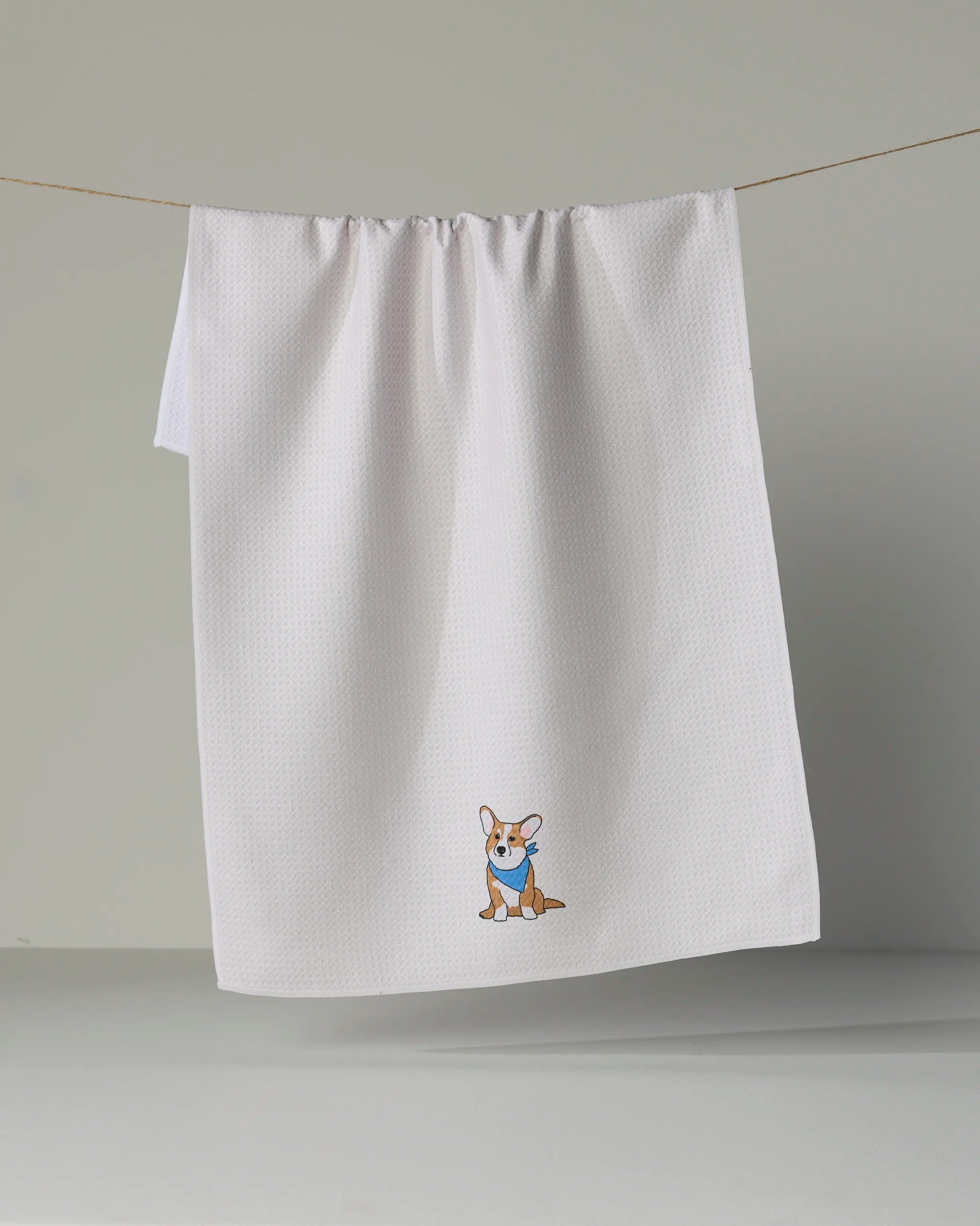 A white kitchen towel from Geometry, called “Corgi Charm,” featuring a small embroidered cartoon corgi wearing a blue scarf, is shown hanging on a clothesline against a plain gray background.