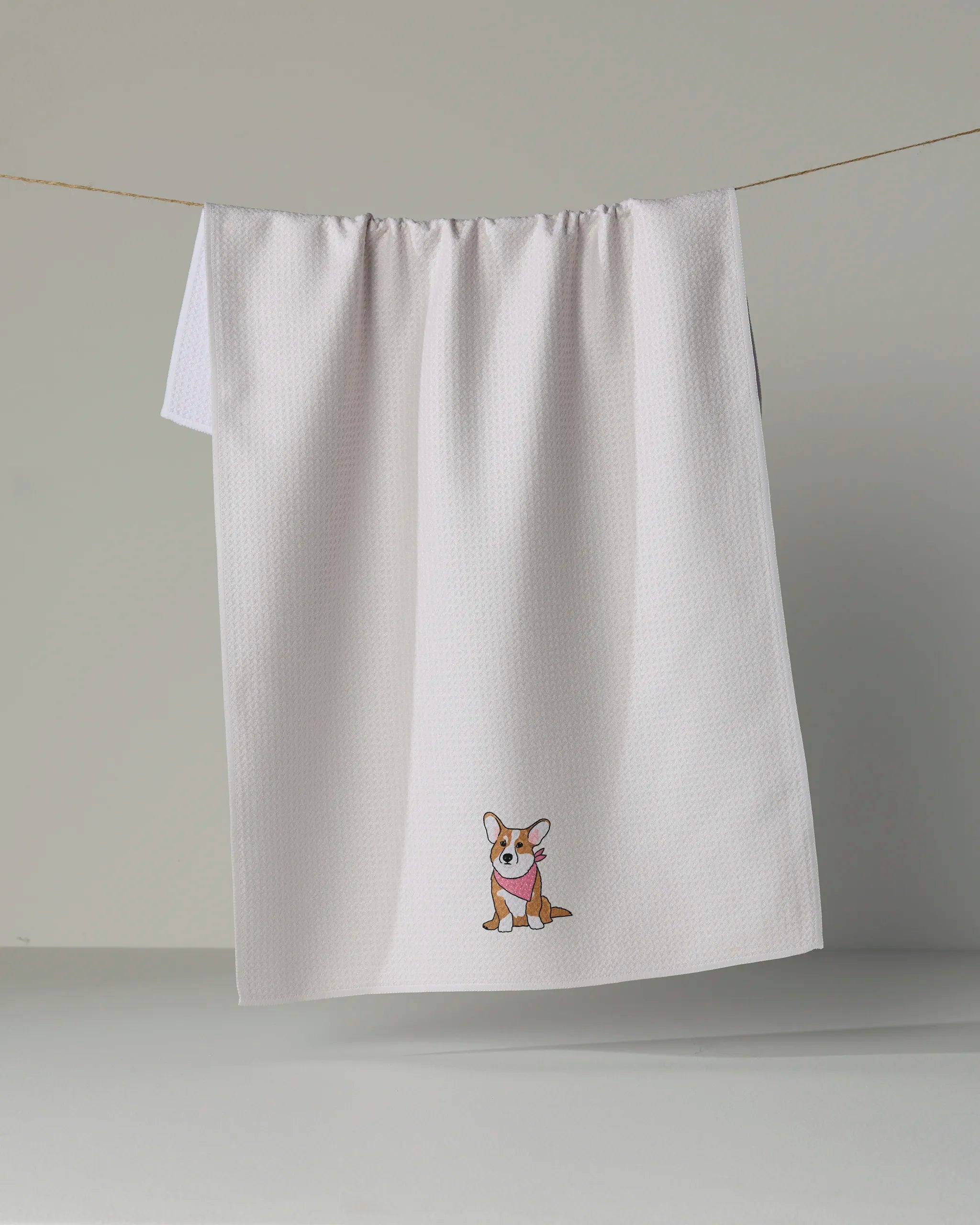 A white kitchen towel from Geometry, called “Corgi Charm,” hangs on a line against a plain background, featuring a small embroidered cartoon corgi near the bottom center.