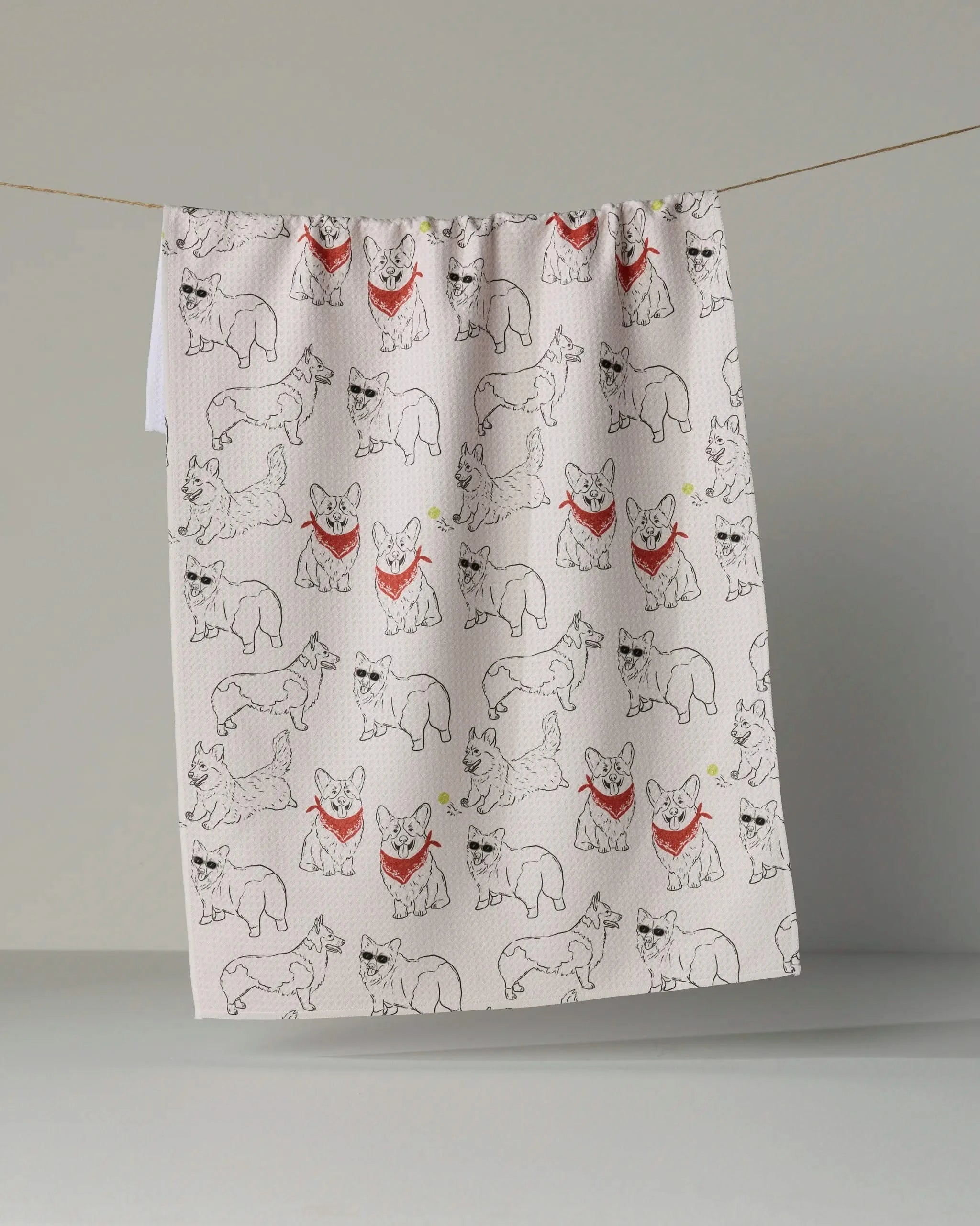 The “Corgi Club” tea towel by Geometry features a repeating pattern of sketched corgis, some sporting red bandanas, and is shown hanging on a string against a plain gray background.
