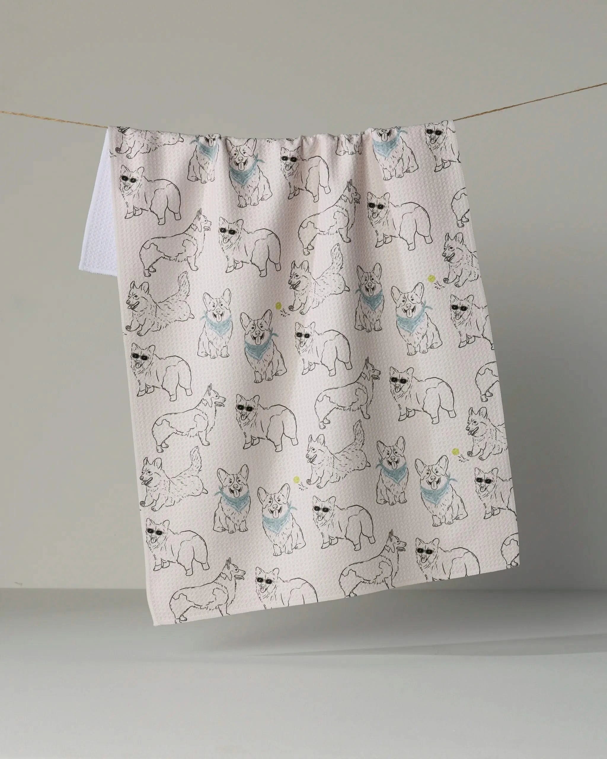 A recycled kitchen towel from Geometry’s Corgi Club collection, featuring a pattern of line-drawn corgis in various poses—some accented with light blue and yellow—hangs on a thin string against a plain light gray background.