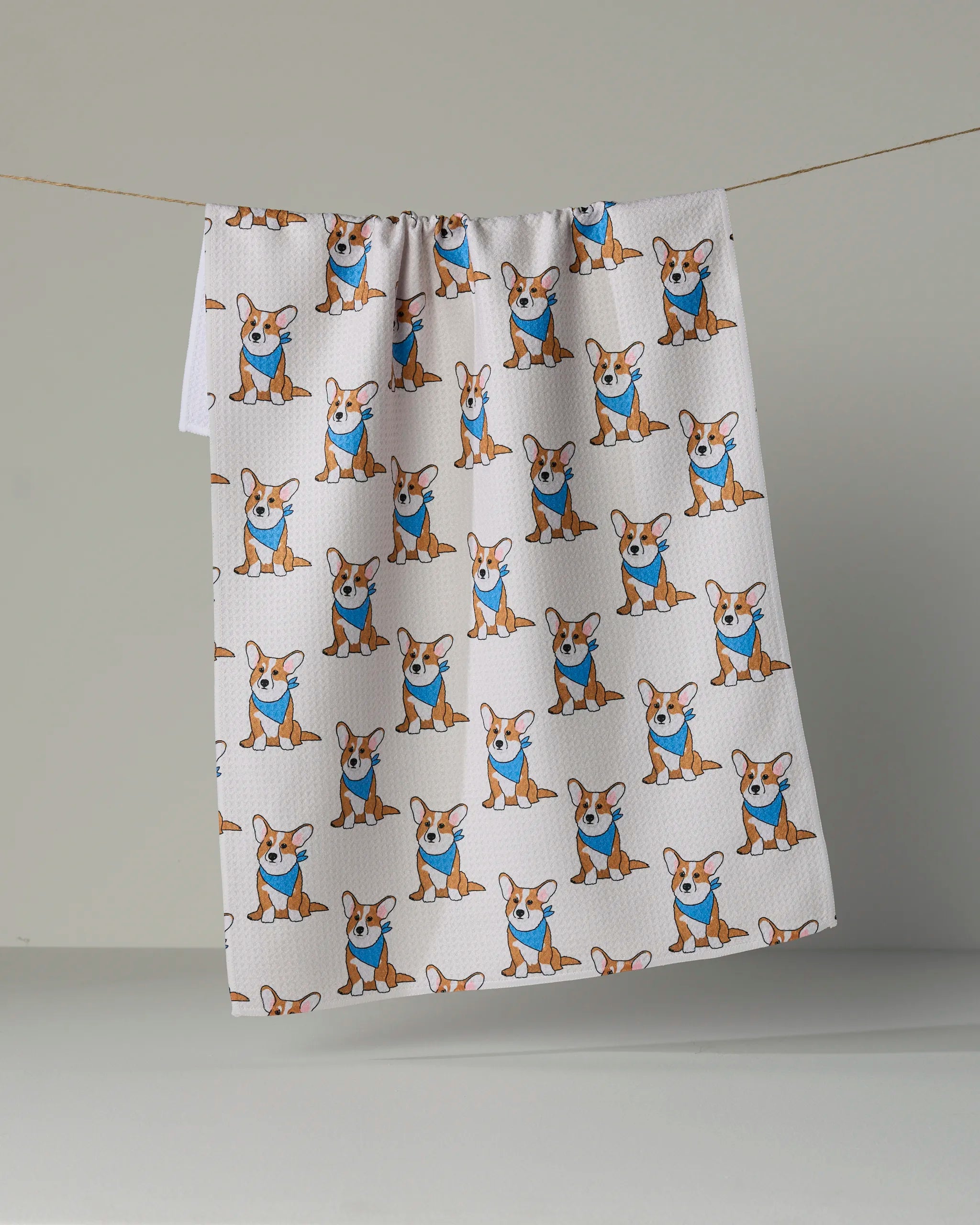 The Geometry “Corgi Pals” kitchen tea towel, crafted from recycled materials, features a charming repeating pattern of cartoon corgis in blue shirts on a crisp white background. Ultra-absorbent and eco-friendly, this stylish towel brings playful personality to your kitchen decor.