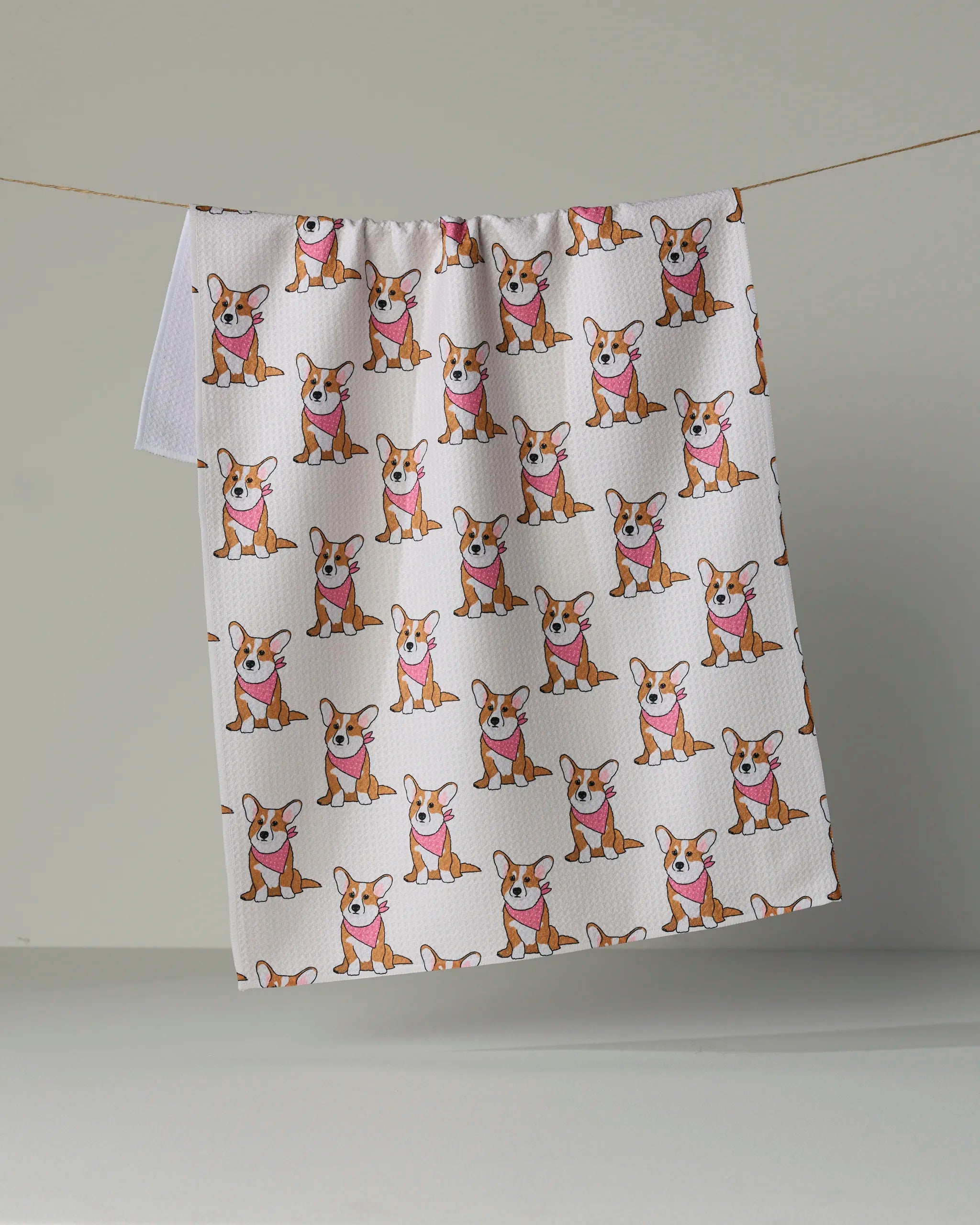Introducing the Corgi Pals kitchen tea towel by Geometry—a charming, ultra-absorbent white towel adorned with a playful repeating pattern of cartoon corgi dogs sporting pink bandanas. Crafted from recycled materials, this adorable and practical towel brightens up your daily routine as it hangs stylishly on a clothesline against a plain background.