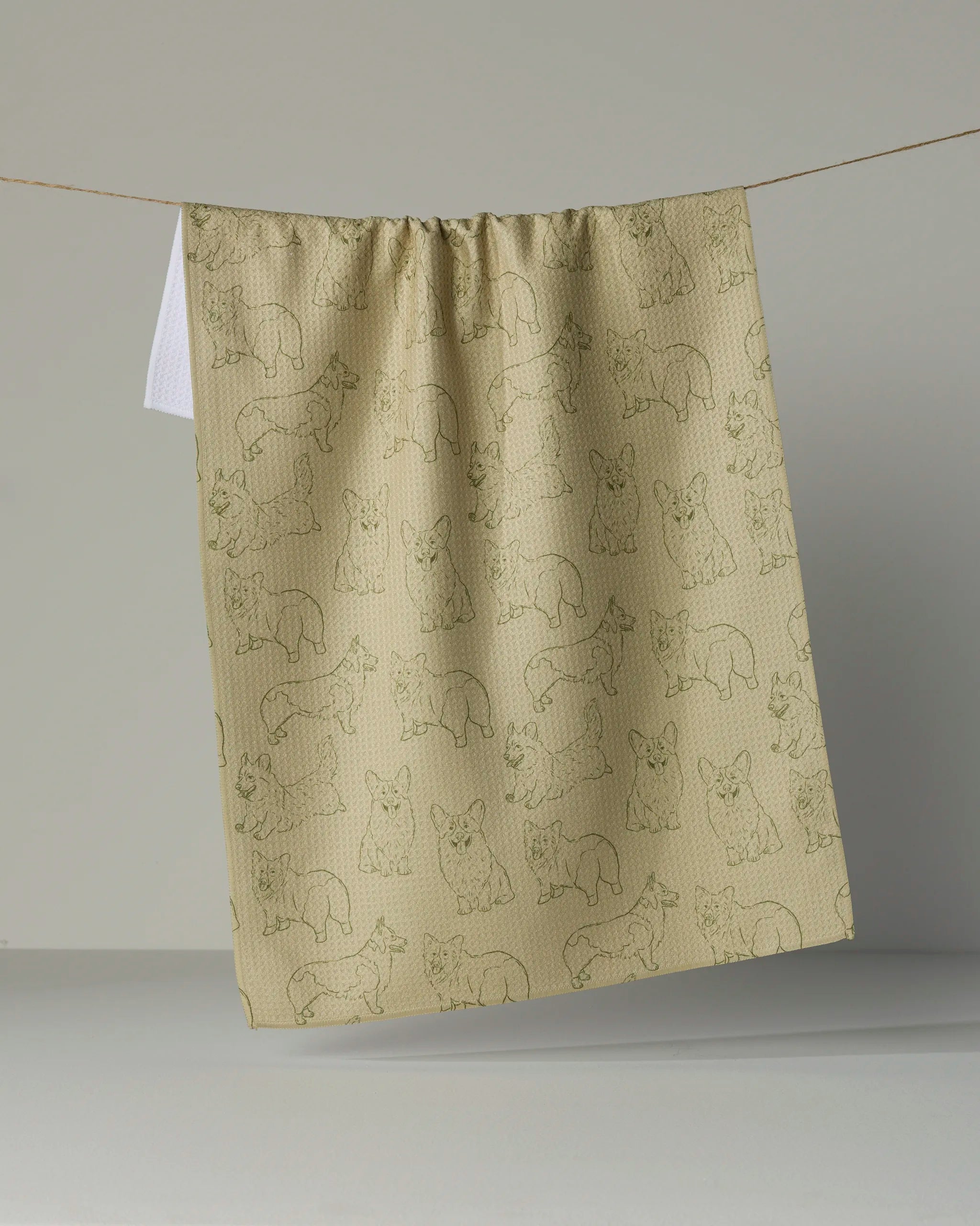 The “Corgi Pups” towel by Geometry features playful green line drawings of corgi puppies on beige fabric, hanging charmingly like a cloth or towel against a plain background—this quick-drying towel effortlessly combines practicality with whimsical appeal for your space.