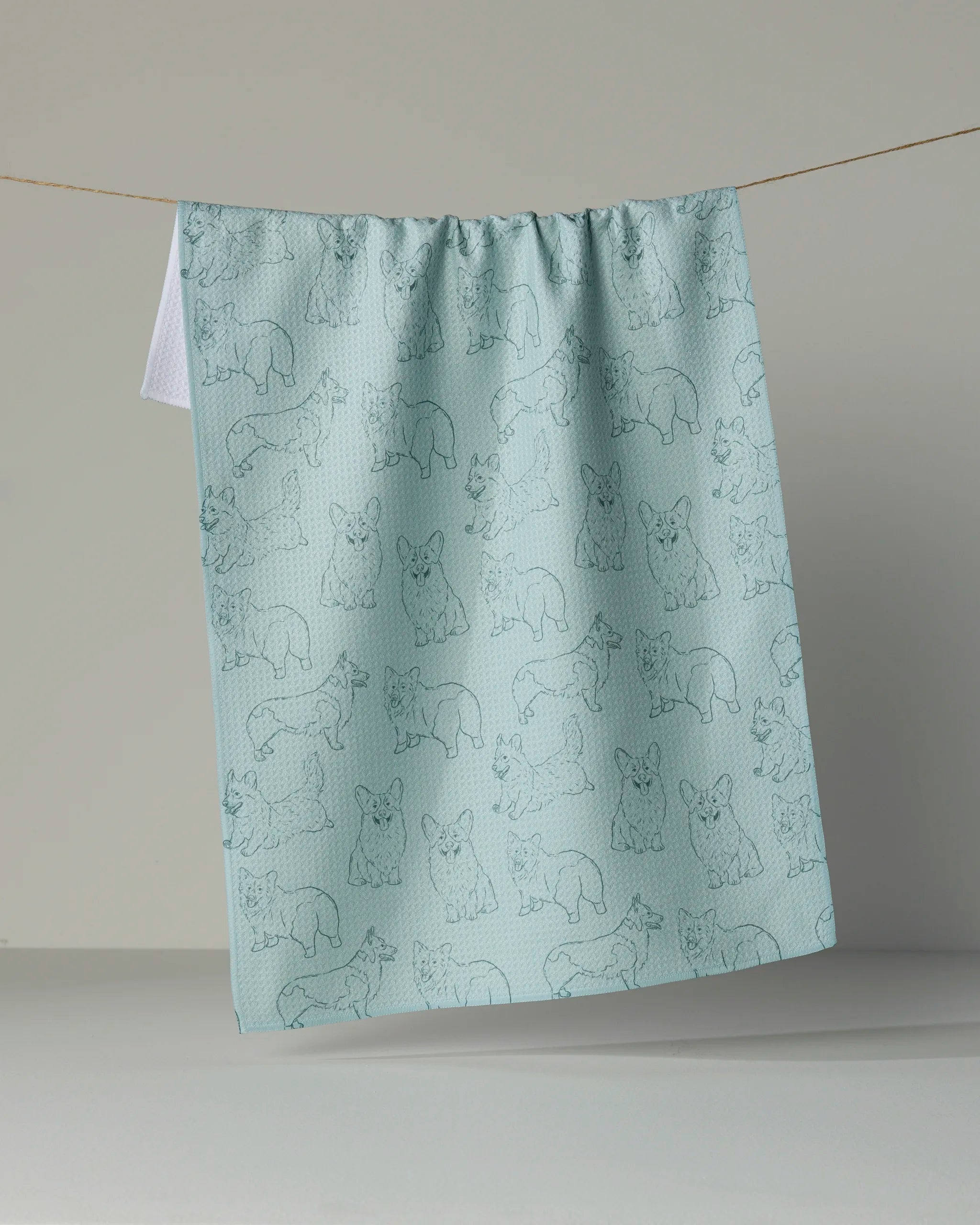 A light blue recycled kitchen towel from Geometry, featuring line drawings of various corgi pups, hangs on a clothesline against a neutral background.