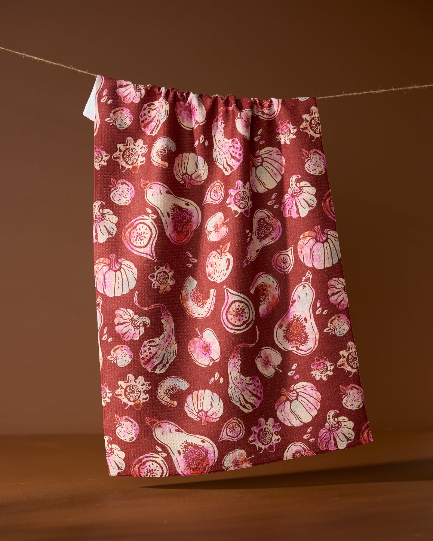 The Geometry Cosecha tea towel, ultra-absorbent and maroon with a pink and white fig and fruit pattern, hangs on a clothesline against a brown background.