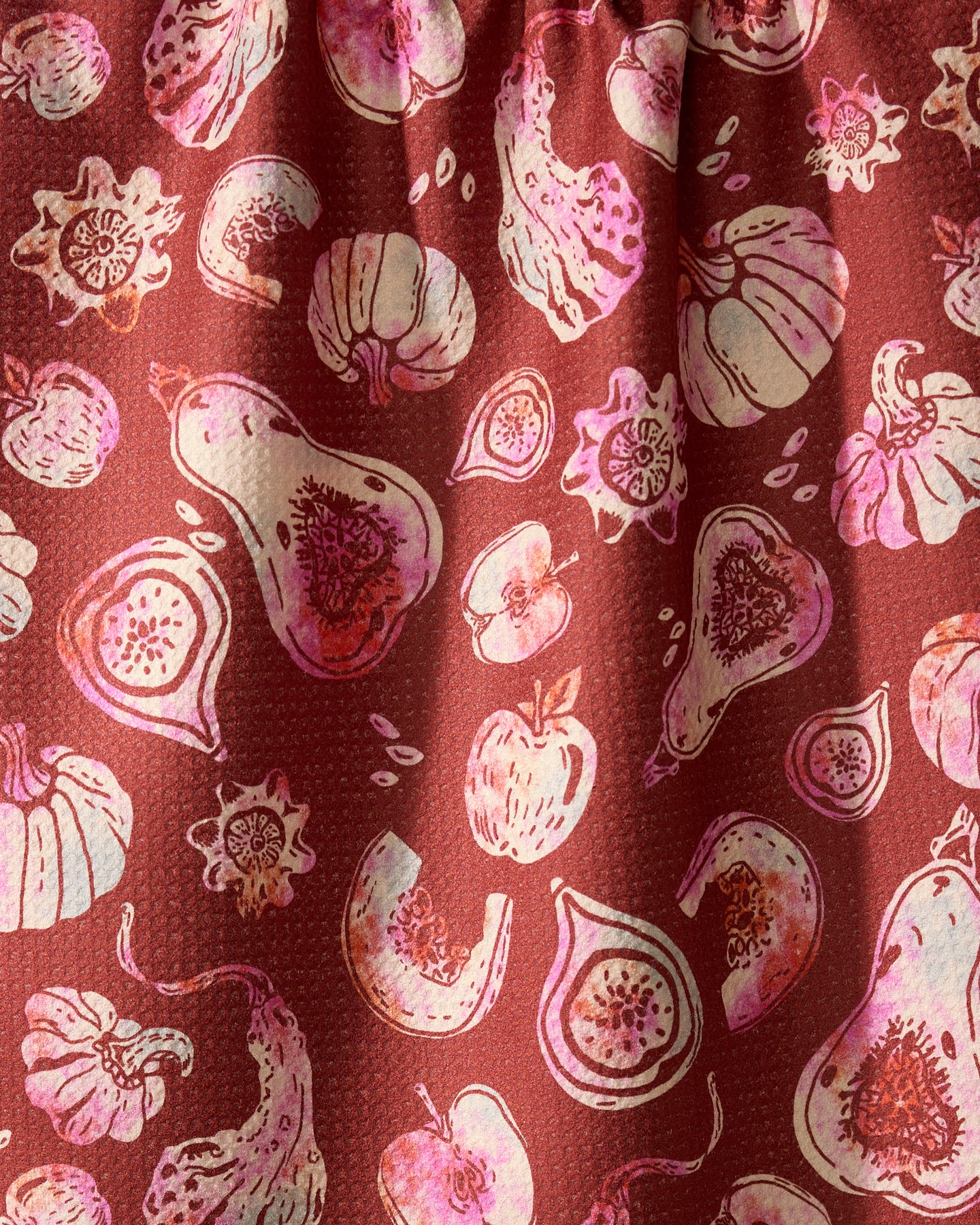 Geometry’s Cosecha recycled kitchen towel features a reddish-brown background with stylized pink and white apples, pumpkins, figs, and squash. Subtle folds and shadows add texture to this eco-friendly tea towel.