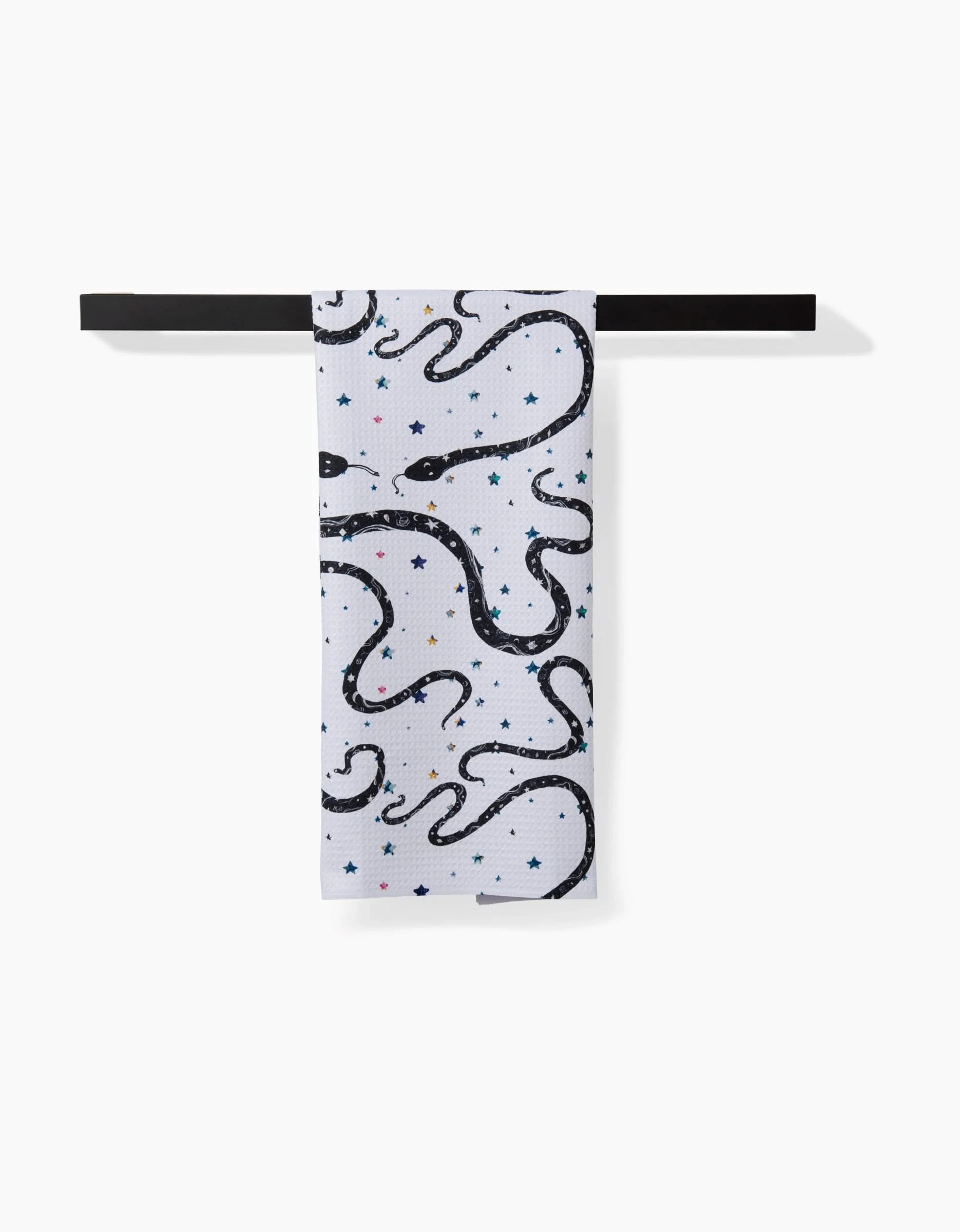 A white ultra-absorbent tea towel, Cosmic Snakes by Pascale Dilger, hangs on a black bar and features black snakes with small colorful stars.