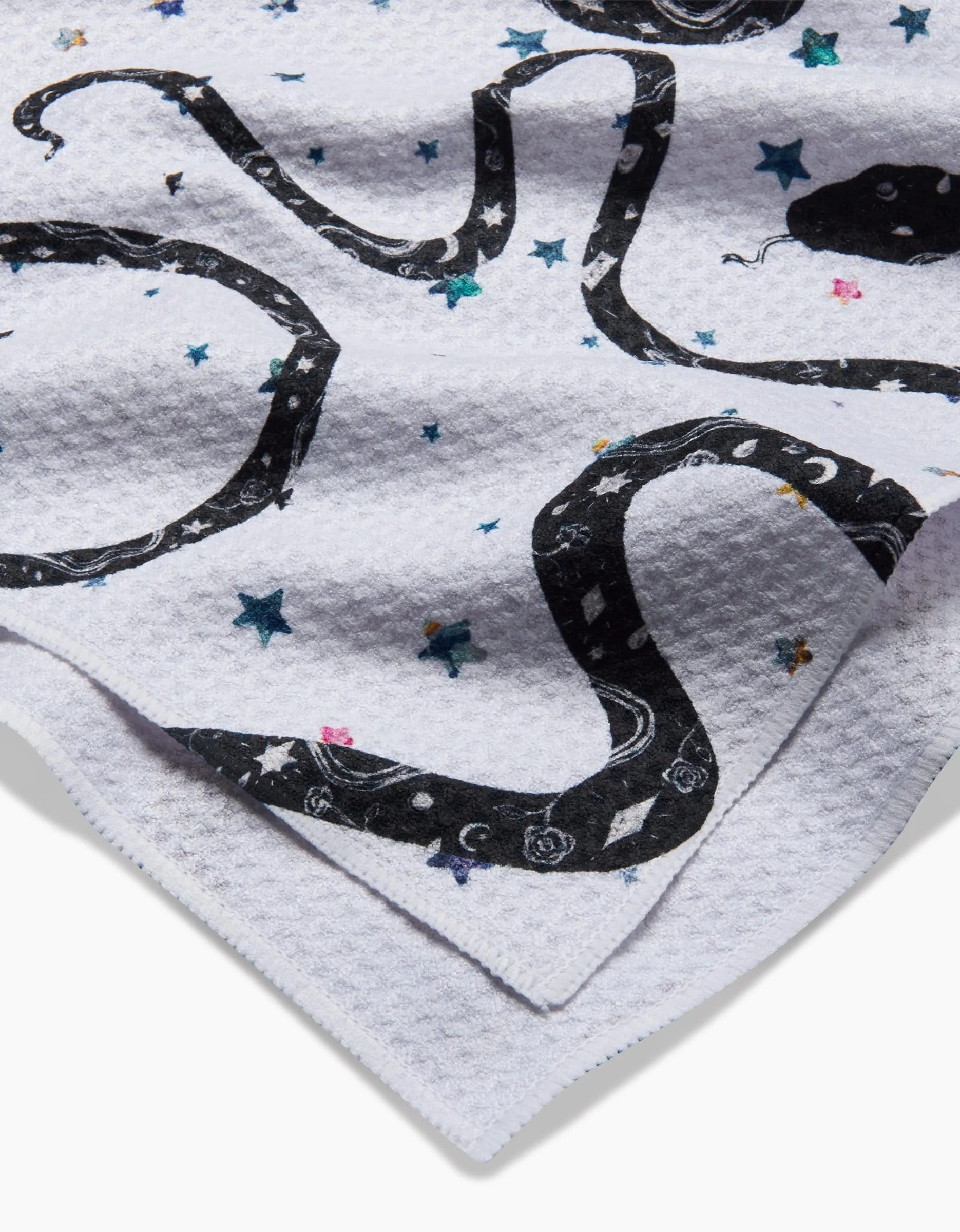 The Cosmic Snakes tea towel by Pascale Dilger features a white textured design with black snake illustrations, stars, and sea motifs on ultra-absorbent recycled materials.