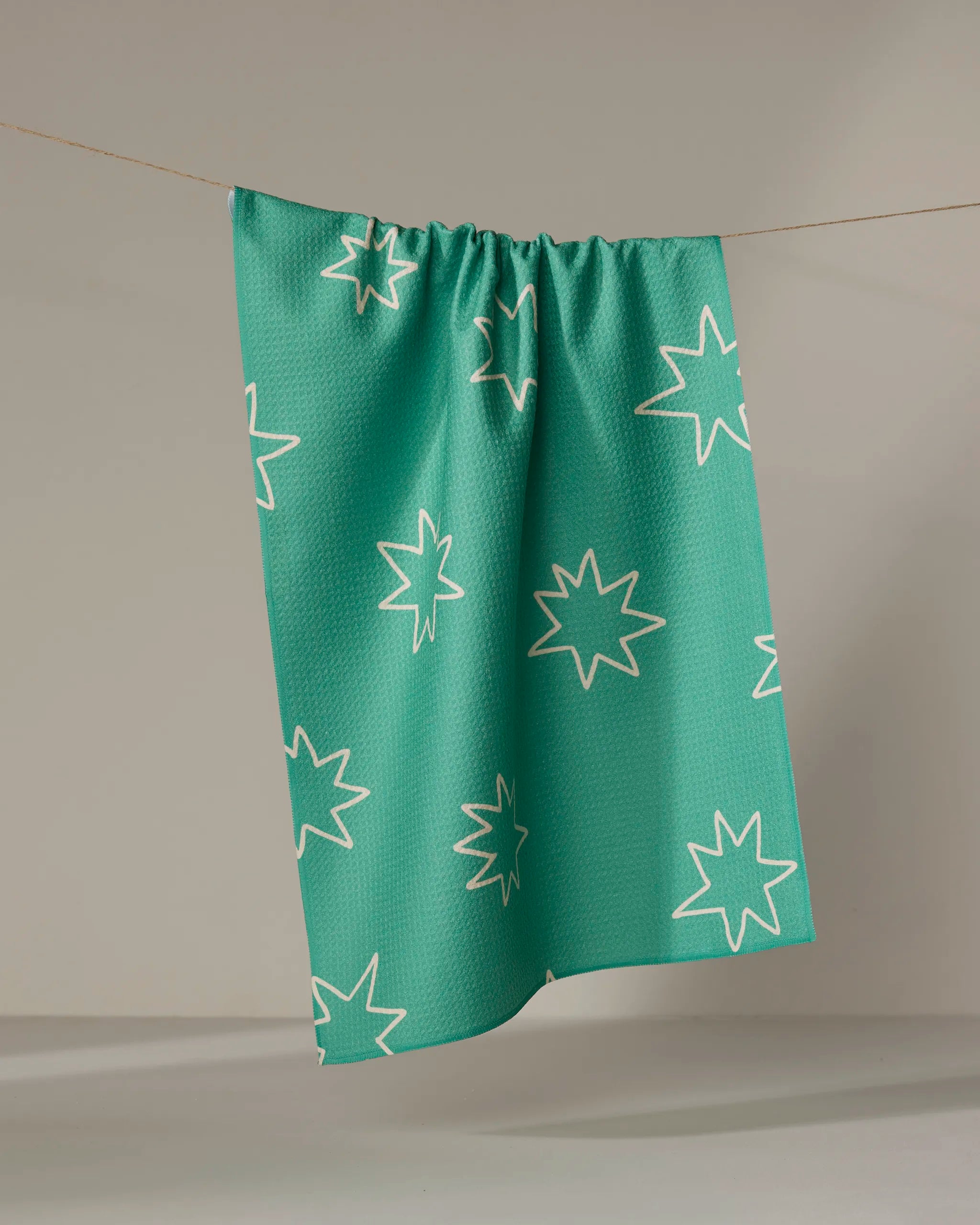 The Beetle Ink Co. Cosmos tea towel, quick-drying in teal with white starburst patterns, hangs from a string against a plain light background.