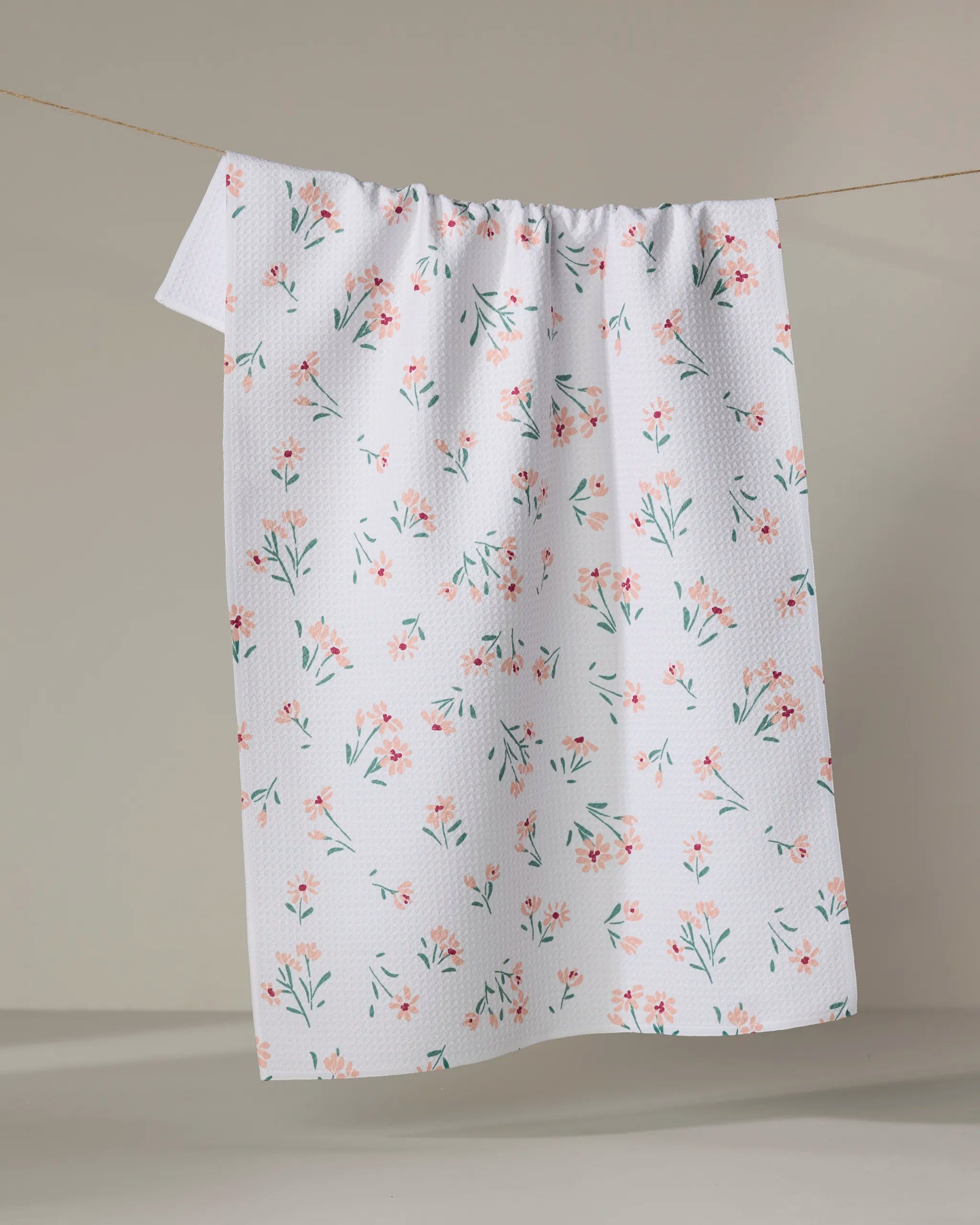 The Cottage Garden tea towel by Hufton Studio, made from recycled materials and featuring a white textured fabric with small pink and green flowers, hangs on a clothesline against a plain beige background.
