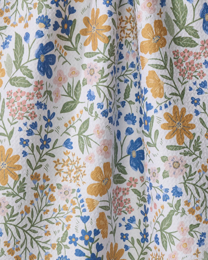 A close-up of Danelys Sidron's “Cottage Meadow Pastel” tea towel, showcasing a soft, eco-friendly fabric with yellow, blue, and pink floral patterns and green leaves on white. Gentle folds highlight its texture.