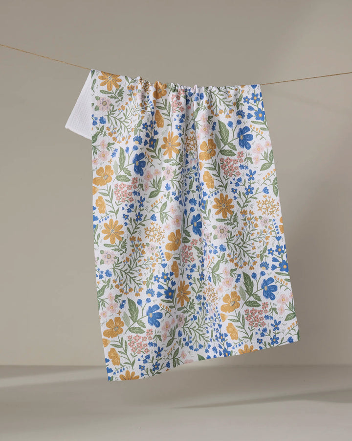 The Danelys Sidron Cottage Meadow Pastel kitchen tea towel, featuring a blue, yellow, pink, and green floral pattern, hangs on a clothesline against a plain beige background.