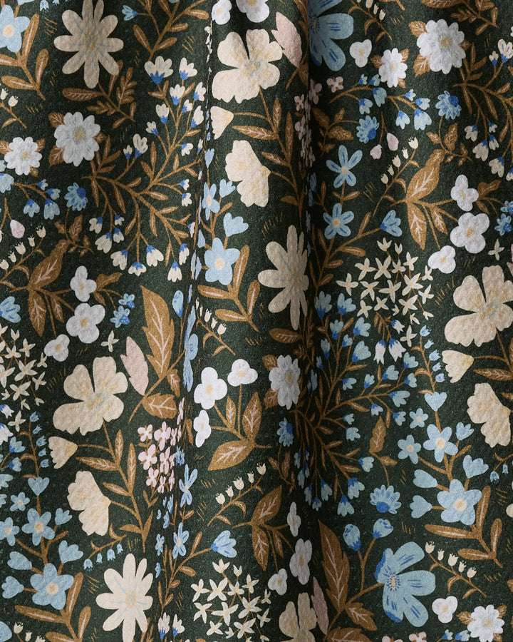 A close-up of the Cottage Meadow Sepia dish towel by Danelys Sidron, featuring an absorbent fabric with white, beige, and blue floral pattern, green leaves, and brown stems on a dark green background with soft folds.