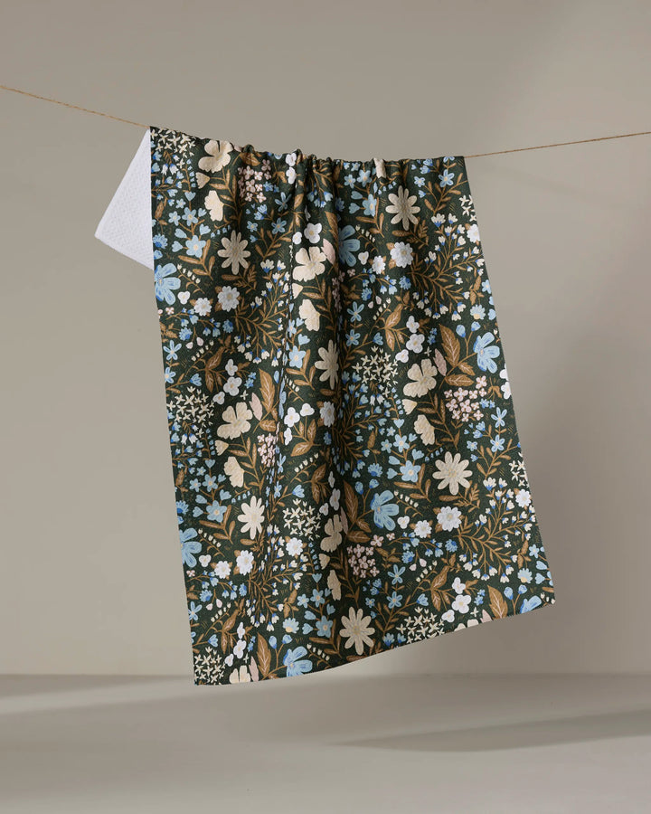 The Danelys Sidron Cottage Meadow Sepia tea towel, featuring blue, white, and brown dark floral tones, hangs on a clothesline against a neutral backdrop with its soft, absorbent fabric visible at one corner.