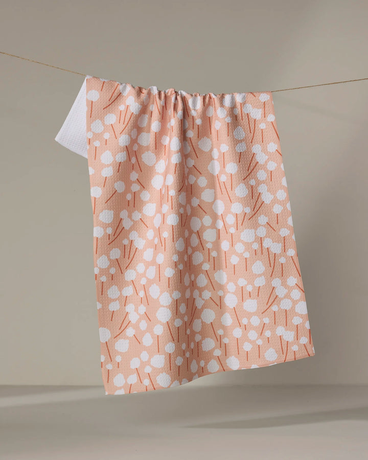 The Coit Creative Cotton Floral towel, featuring a peach color with white abstract dots and lines, hangs on a clothesline against a neutral background.