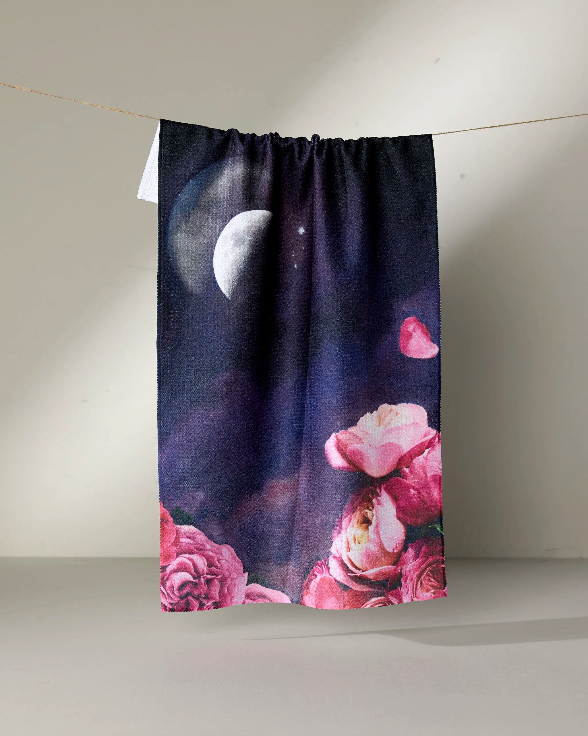 The Counterglow kitchen tea towel by Stefanie Bales hangs on a line, displaying a crescent moon and stars over pink roses. Soft shadows and a neutral backdrop highlight this ultra-absorbent towel crafted from recycled materials.