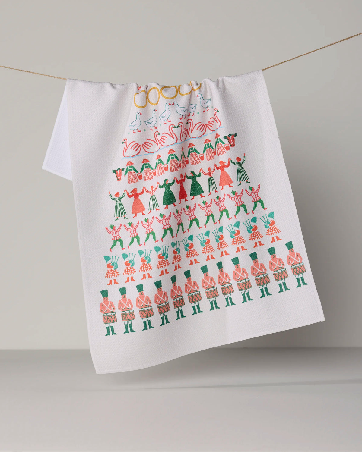 The “Counting Christmas” tea towel by Geometry hangs on a line, adorned with vibrant illustrations of people stacked in a festive Christmas tree shape and dressed in red, green, and pink attire. Crafted from quick-drying recycled materials, it brings holiday cheer and sustainability to your kitchen.