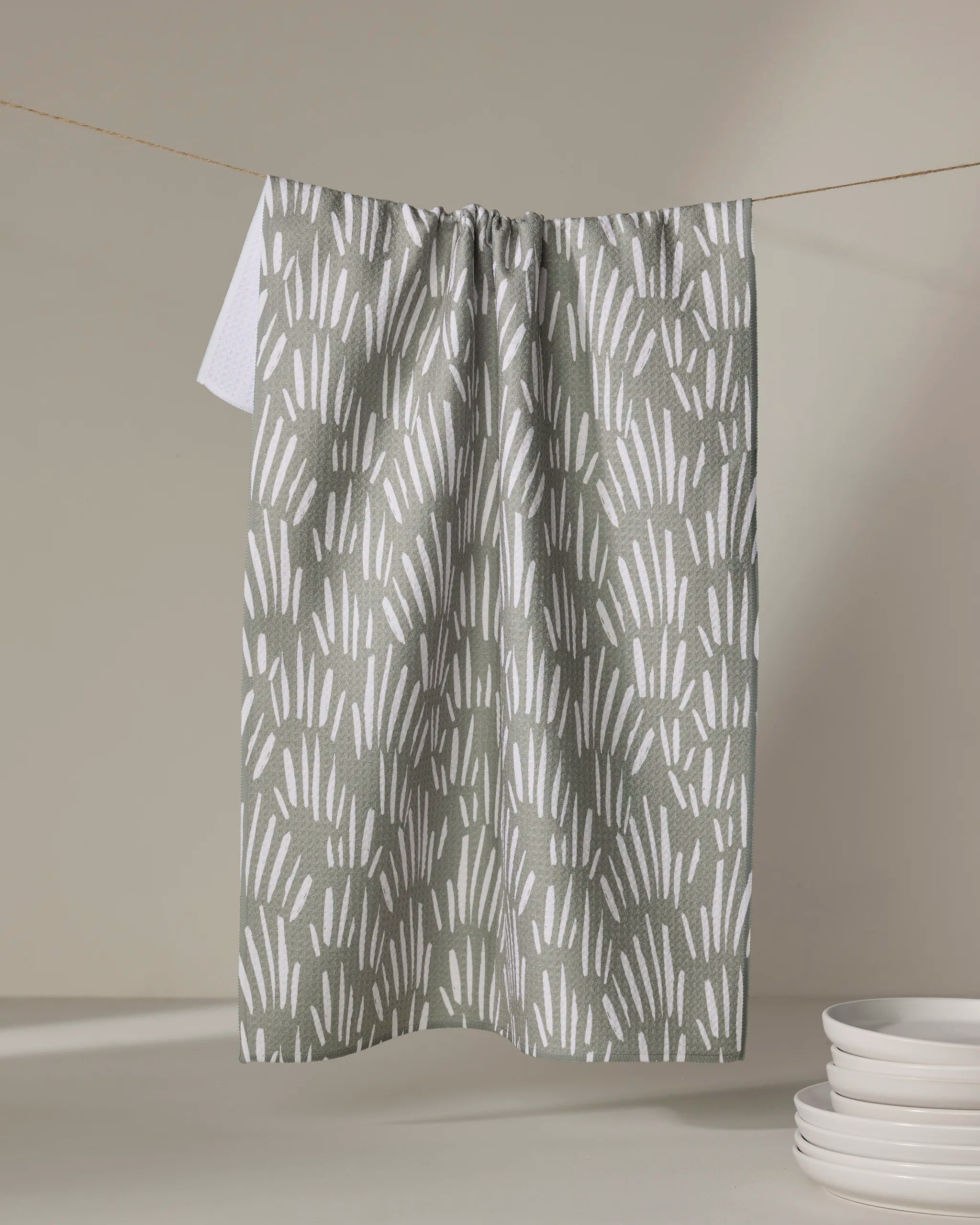 A Hufton Studio "Country Fields" green and white patterned kitchen towel hangs on a clothesline, with a stack of white plates nearby on a light surface.