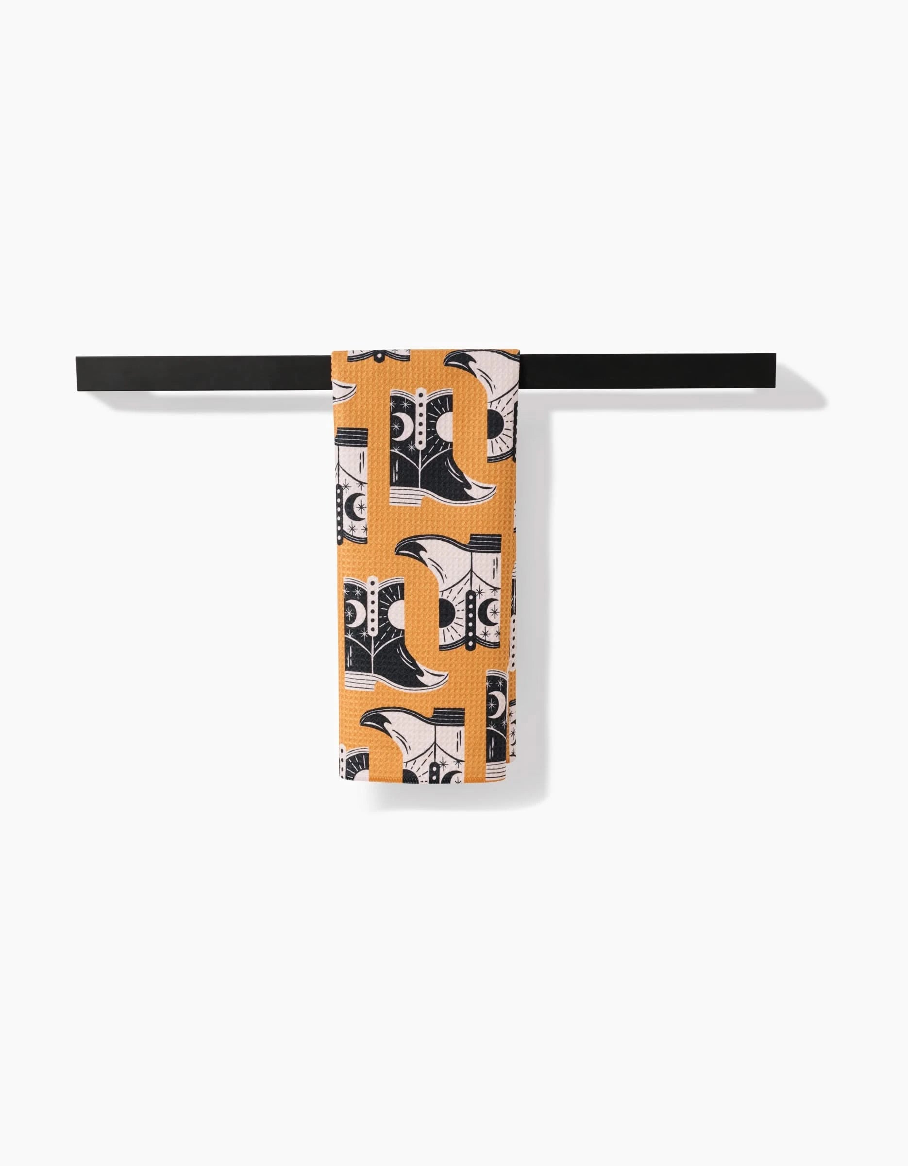 A Megan Roy Cowboy Boots tea towel featuring orange, black, and white abstract boot designs hangs neatly folded over a simple horizontal black towel bar, highlighting its ultra-absorbent Geoweave fabric against a clean white backdrop.