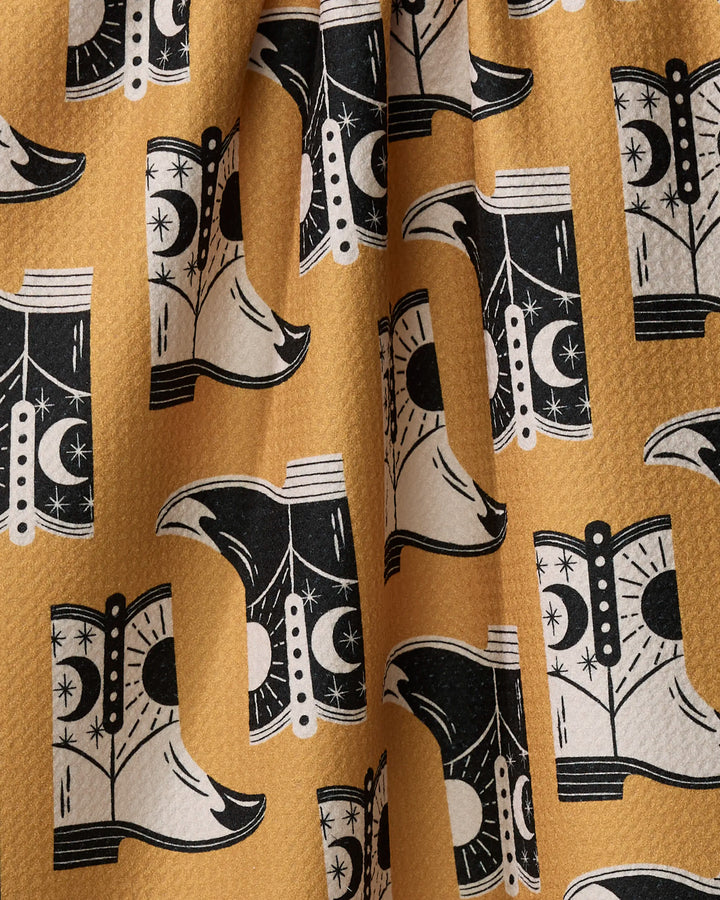 A close-up of Megan Roy’s "Cowboy Boots" quick-drying yellow fabric, featuring a repeating black and white cowboy boot pattern with moon and star motifs. The material is slightly wrinkled.