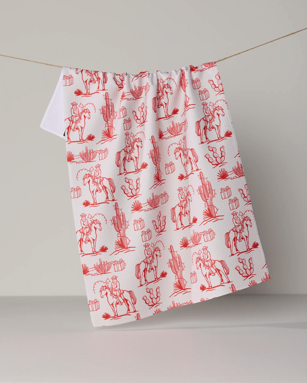 Geometry’s Cowboy Claus tea towel, crafted from recycled materials, features red illustrations of cowboys on horseback, cacti, and desert plants against a white background and is pictured hanging on a string in front of a neutral backdrop.
