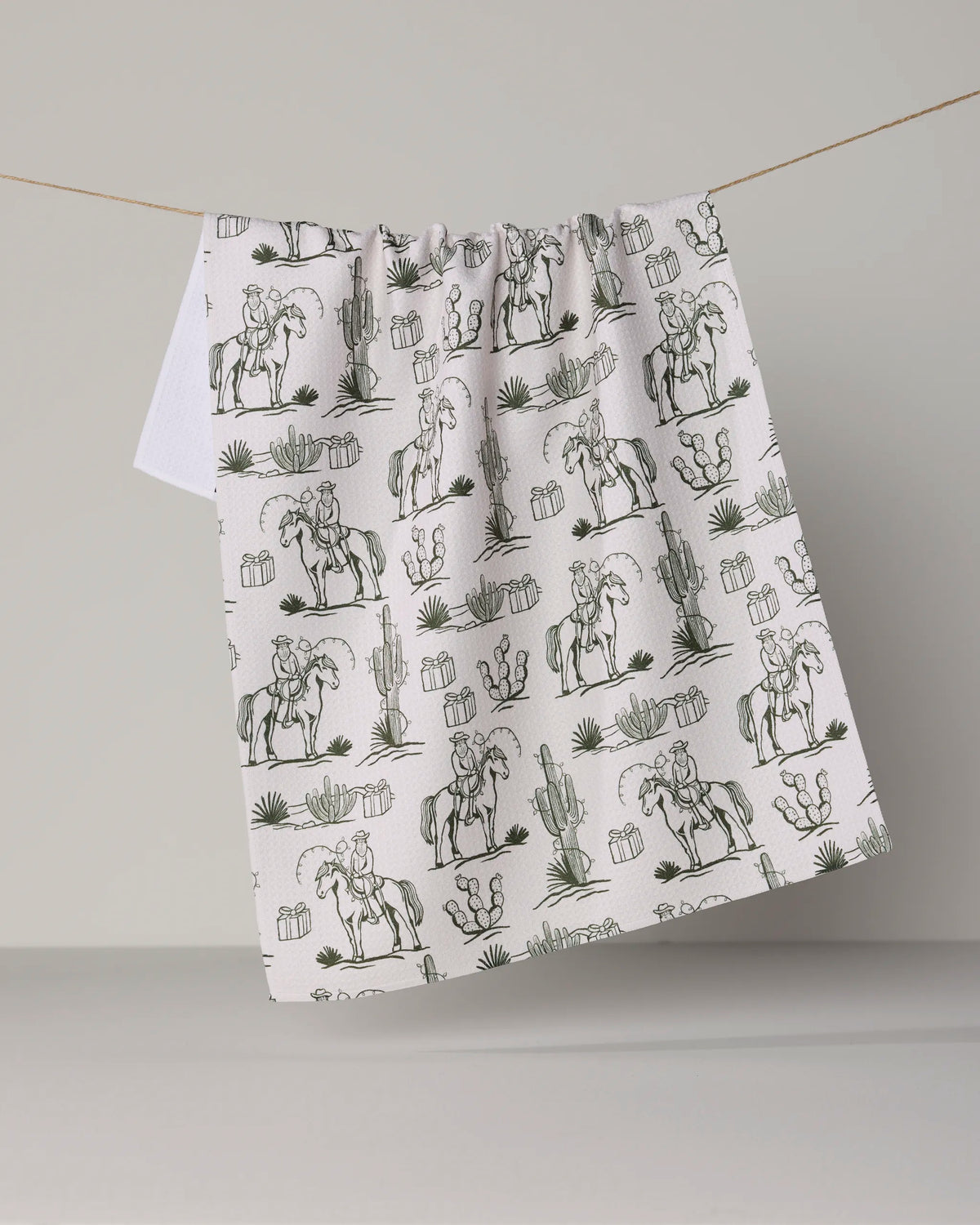 The Geometry “Cowboy Claus” kitchen towel, made from recycled materials, features an ultra-absorbent white fabric adorned with a repeating black line drawing of a cowboy riding a horse among cacti and desert plants, and is shown hanging on a clothesline.