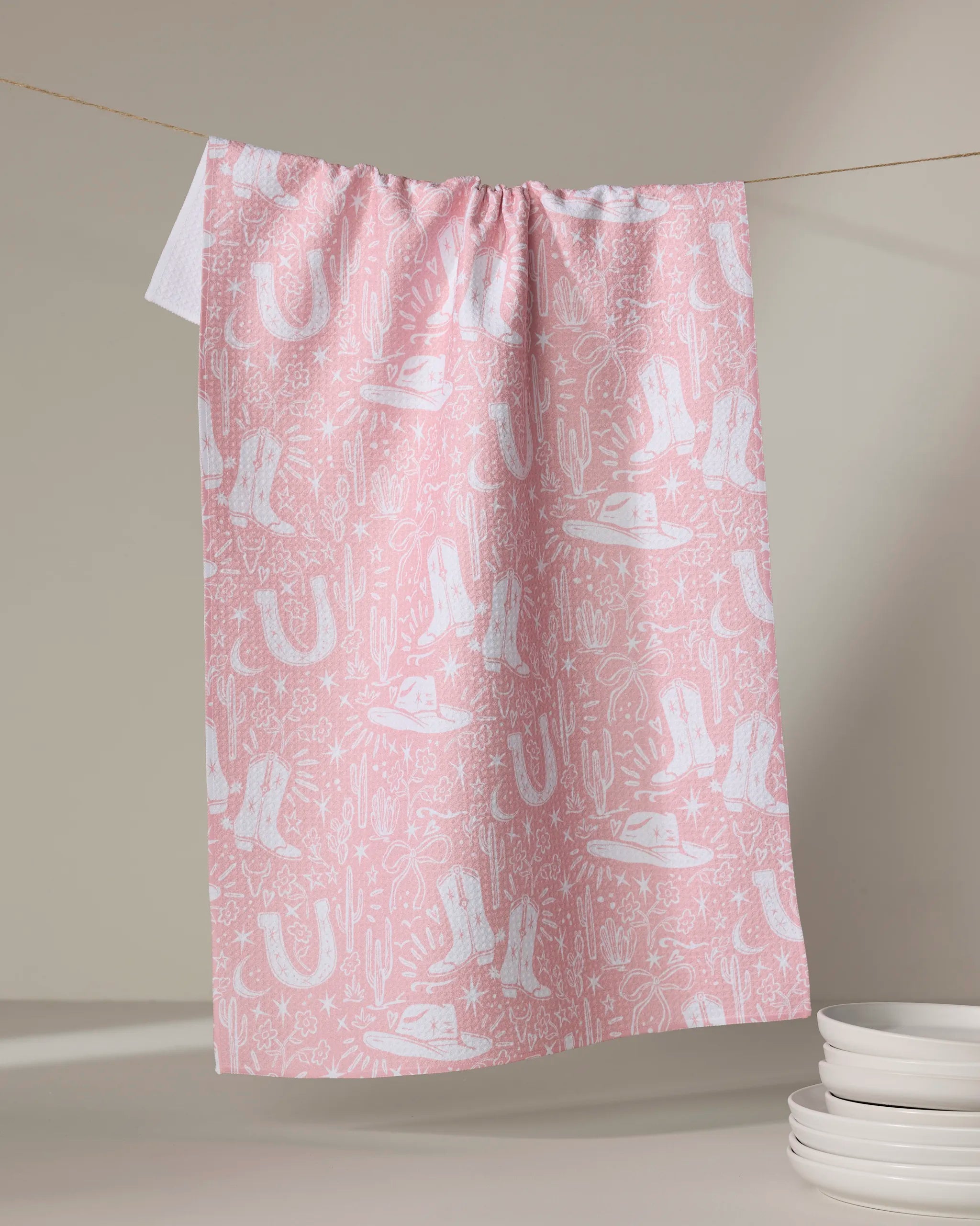 The "Cowgirl Chic" recycled pink kitchen towel by Aubrey Fairchild, featuring white cowboy-themed illustrations, hangs on a clothesline above stacked white plates against a neutral backdrop.