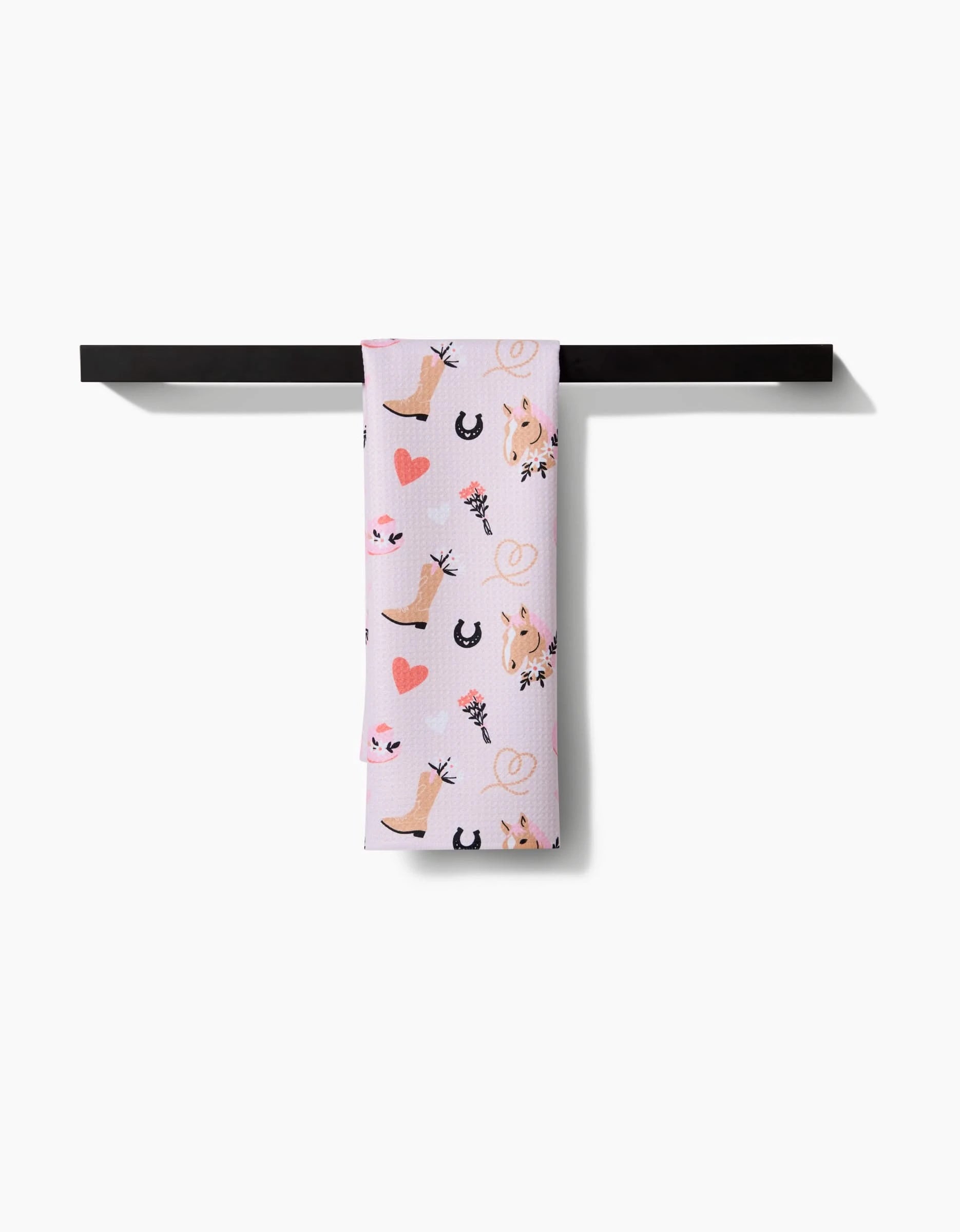 The Cowgirl Love recycled kitchen towel by Amberleigh Adoff, featuring pink hearts, horses, horseshoes, and floral prints, hangs over a black bar against a white background.