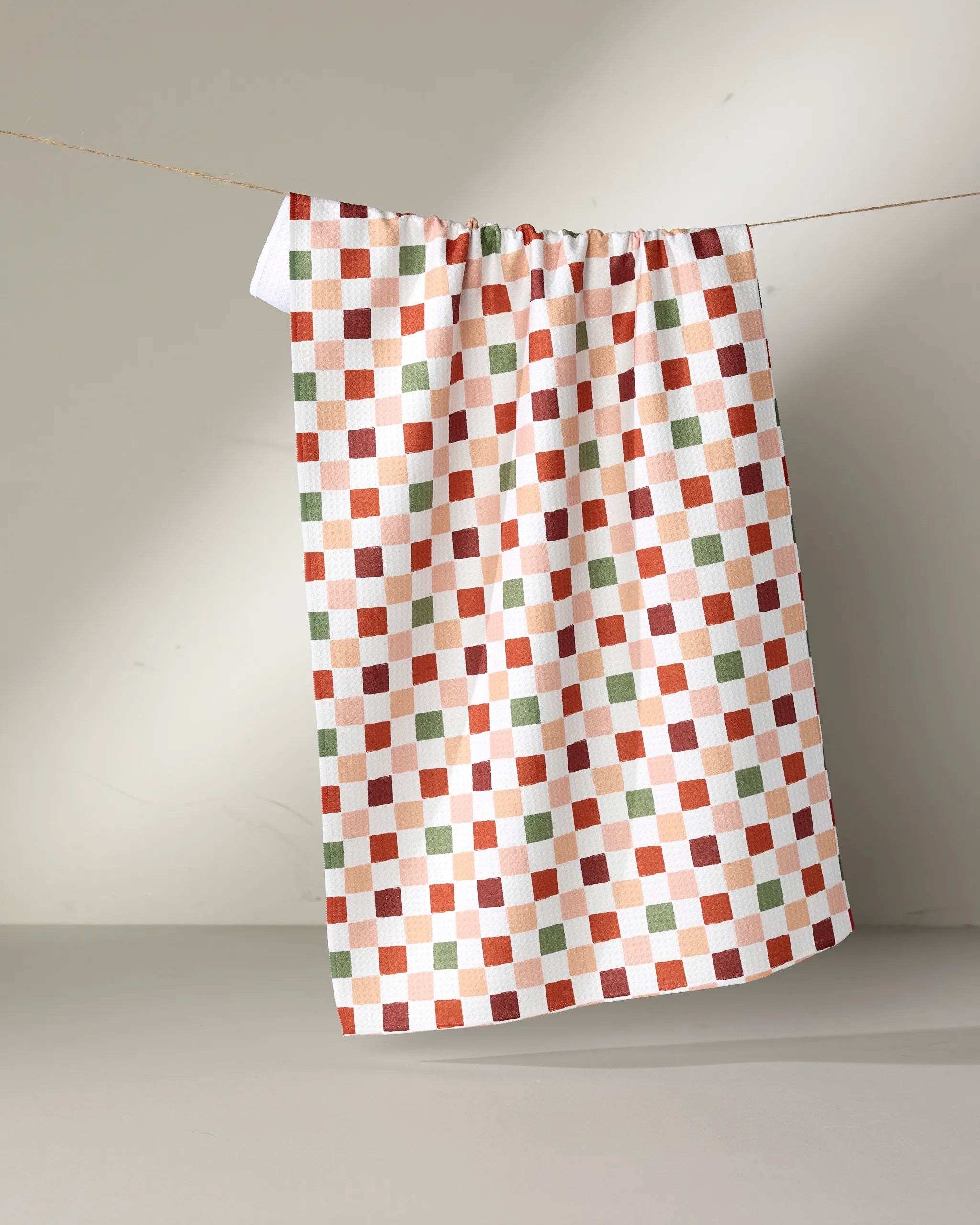 The Ashley Reeves Cozy Check tea towel, featuring a red, green, orange, and white checkered pattern, hangs from a string against a neutral background.