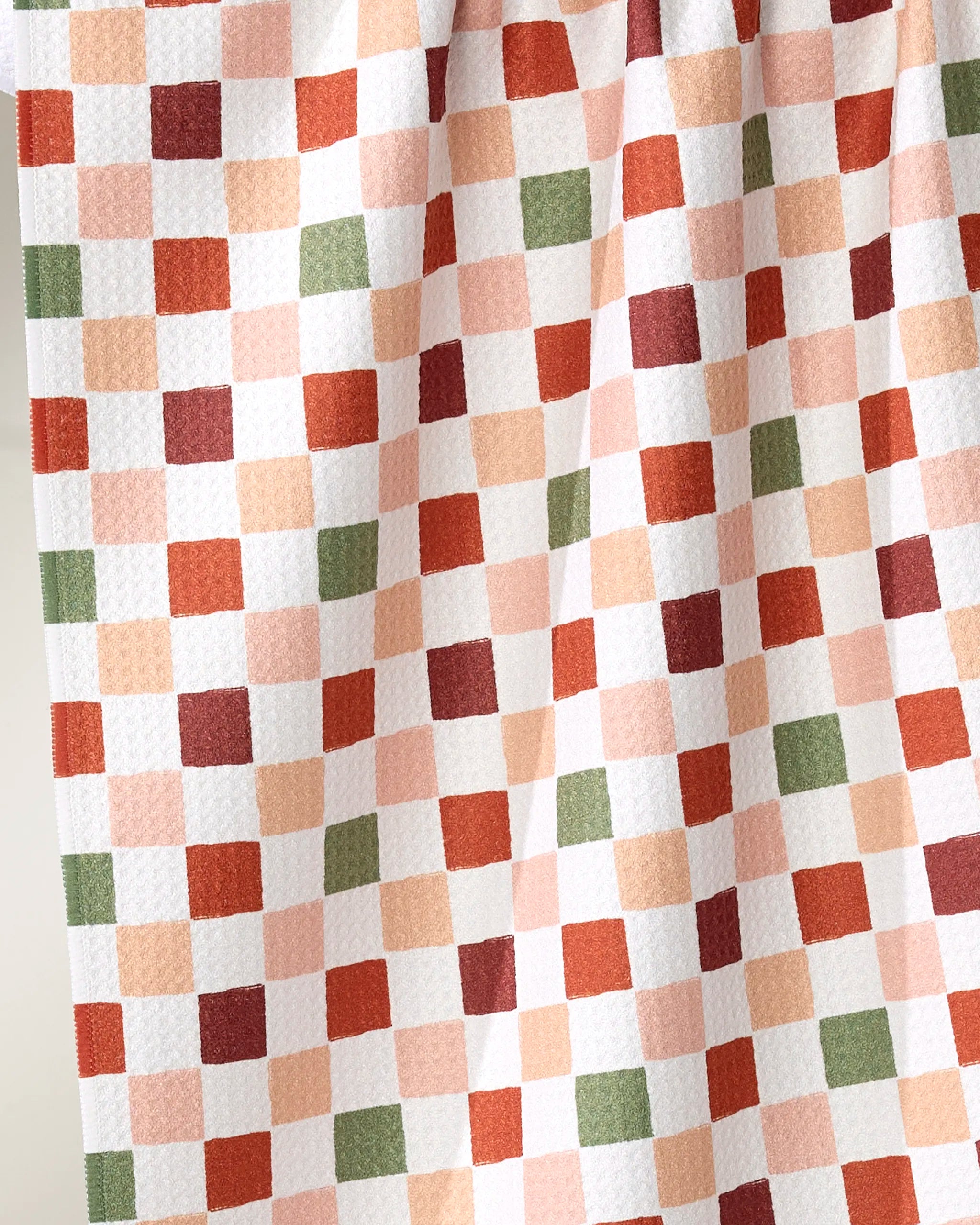 A close-up of Ashley Reeves’ Cozy Check tea towel shows its ultra-absorbent fabric with a checkered pattern of orange, red, green, and peach squares on white, softly gathered at the top.