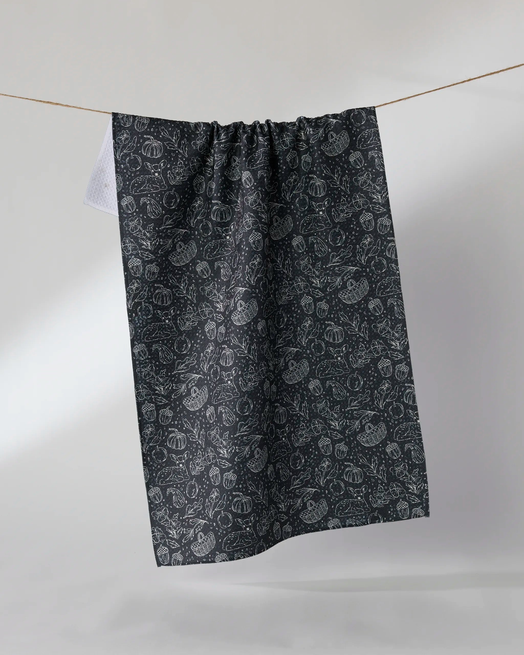 The Geometry Cozy Flora kitchen tea towel, crafted from recycled materials for sustainable style, features a dark hue with an intricate botanical pattern and hangs on a clothesline against a plain light background. Ultra-absorbent and eco-friendly.