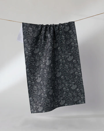 The Geometry Cozy Flora kitchen tea towel, crafted from recycled materials for sustainable style, features a dark hue with an intricate botanical pattern and hangs on a clothesline against a plain light background. Ultra-absorbent and eco-friendly.