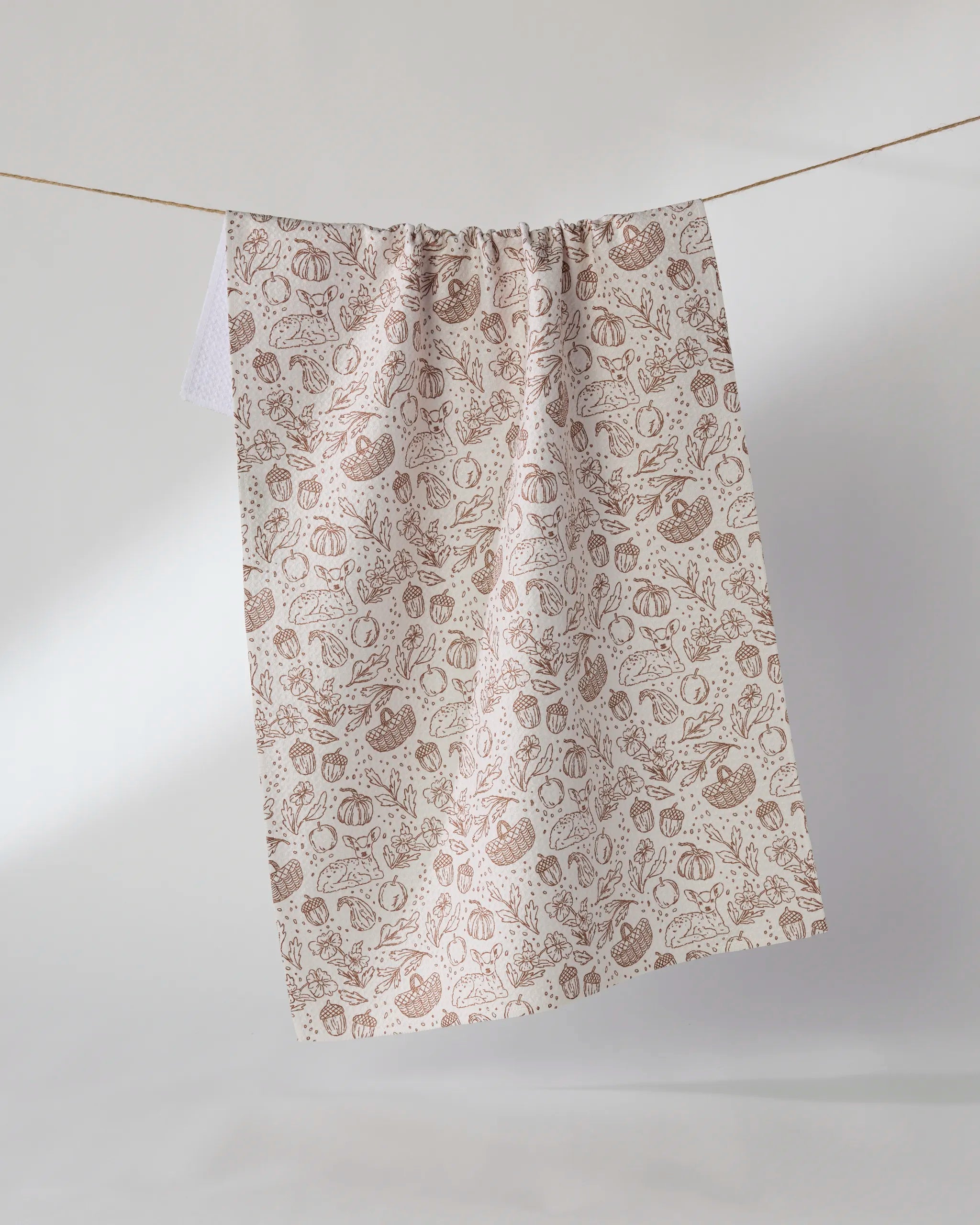 The Cozy Flora tea towel by Geometry, made from recycled materials, displays a brown vegetable line art pattern on beige fabric and hangs against a plain light background.