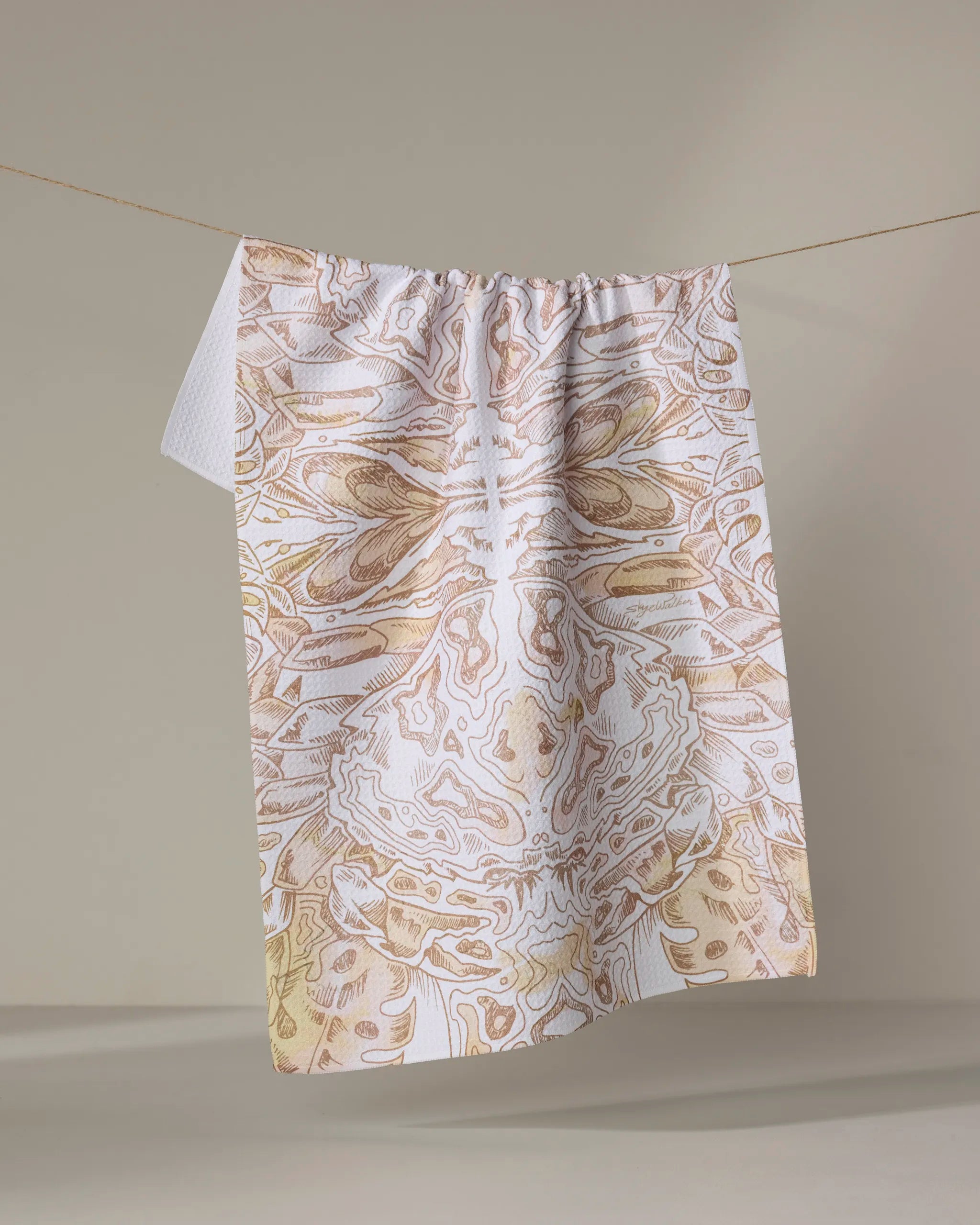 The Skye Walker "Crab" dish towel, featuring abstract symmetrical beige and white patterns, hangs on a clothesline against a neutral background.
