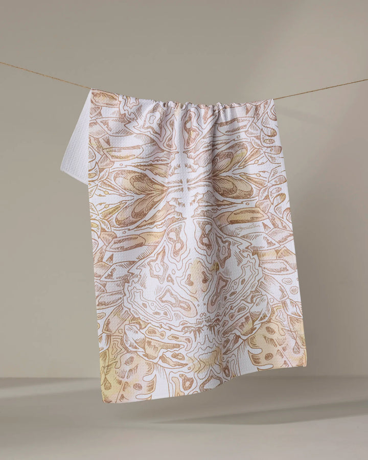 The Skye Walker "Crab" dish towel, featuring abstract symmetrical beige and white patterns, hangs on a clothesline against a neutral background.